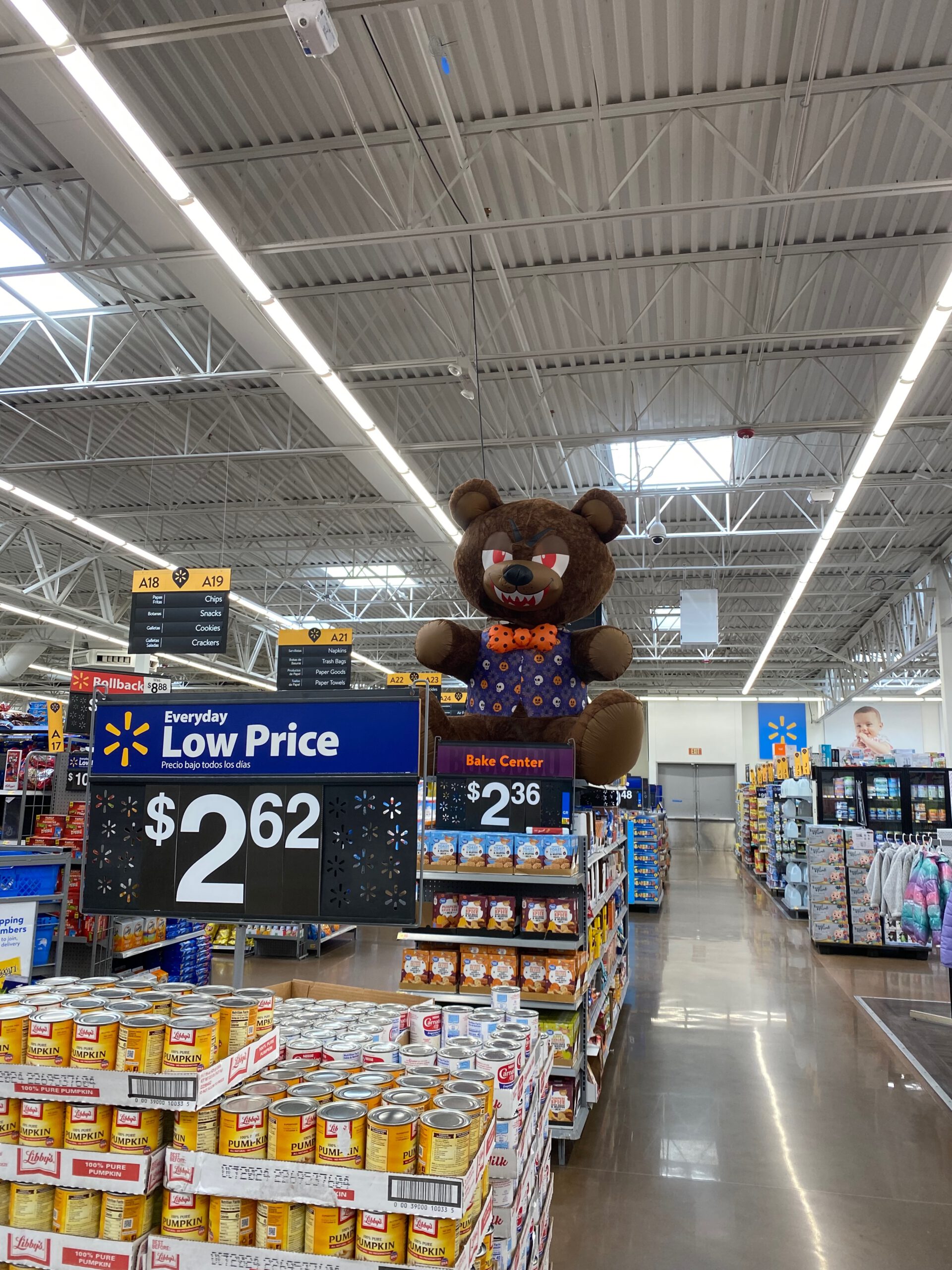 Interior view of Walmart Supercenter Beaumont California showing a Halloween-themed grocery aisle with a giant spooky teddy bear decoration above the bake center, everyday low price signage, canned pumpkin displays, wide aisles, and a high industrial ceiling typical of large American supermarkets.