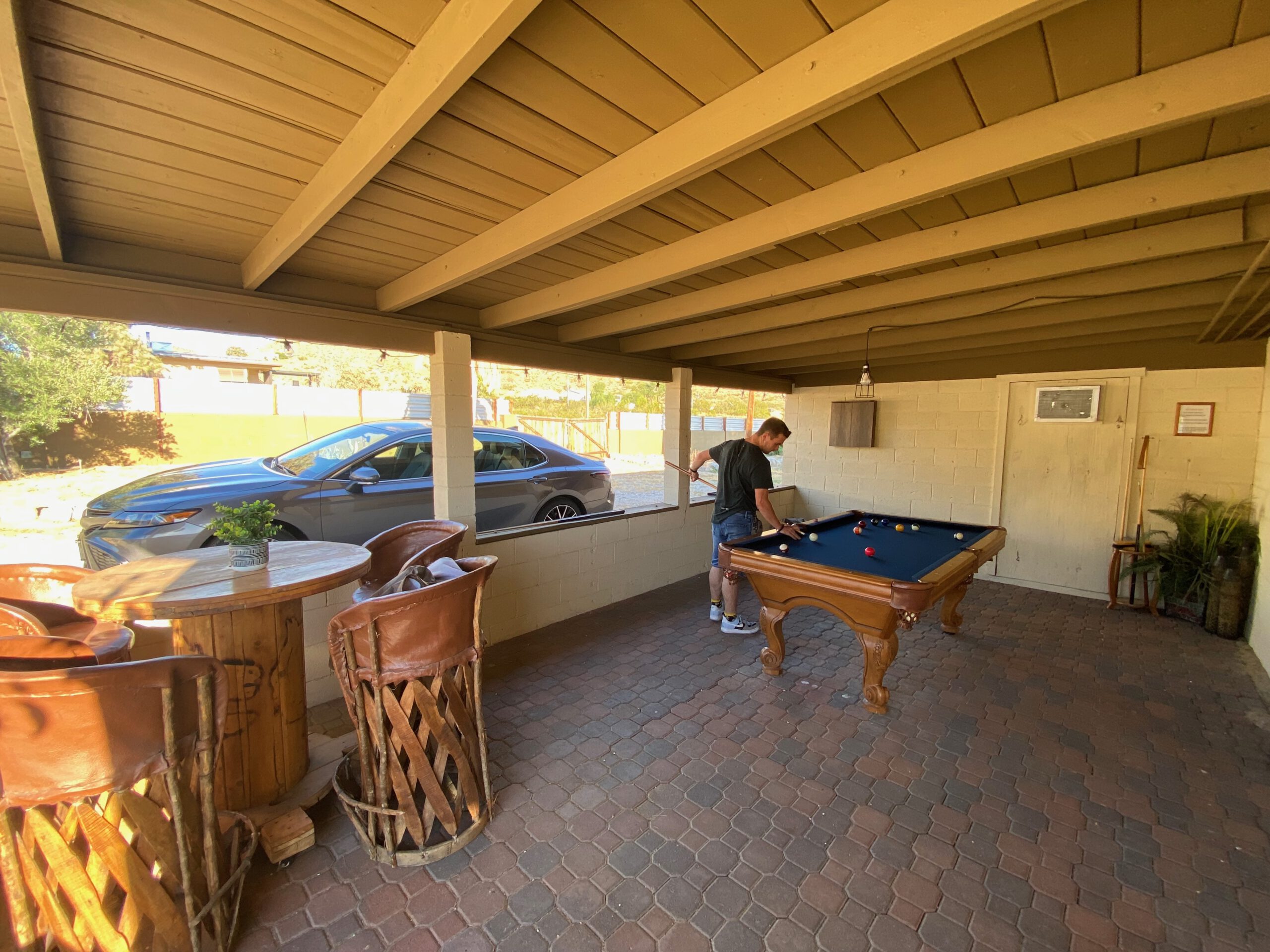 Covered outdoor patio at Casa Rustica Morongo Valley Airbnb featuring a wooden pool table, round seating table with chairs, brick flooring, and a parked car visible in the driveway during a sunny desert afternoon.
