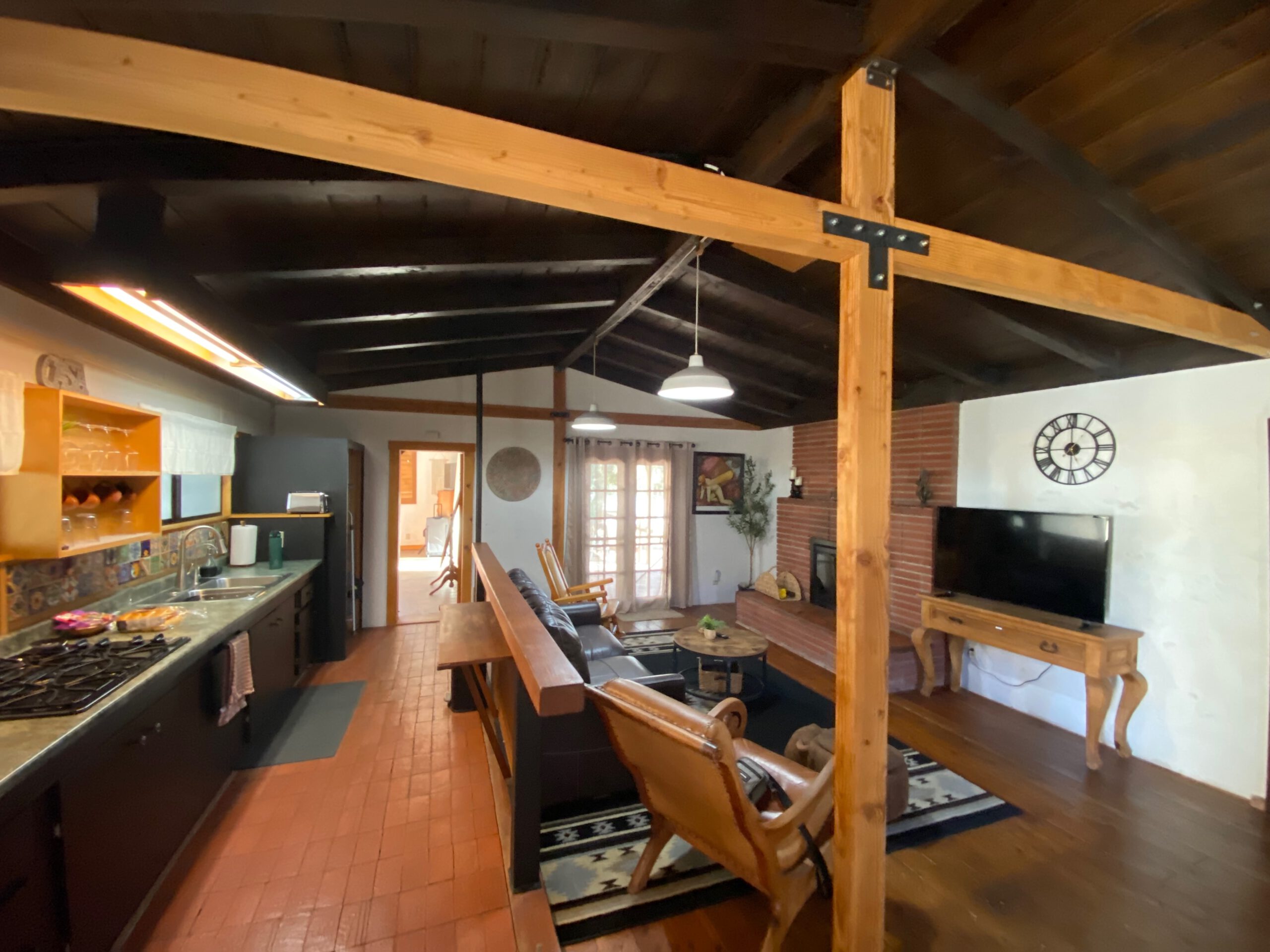 Open-plan living area of Casa Rustica Morongo Valley Airbnb with rustic wooden beams, fully equipped kitchen, bar table, leather seating, brick fireplace, and desert-style interior design.