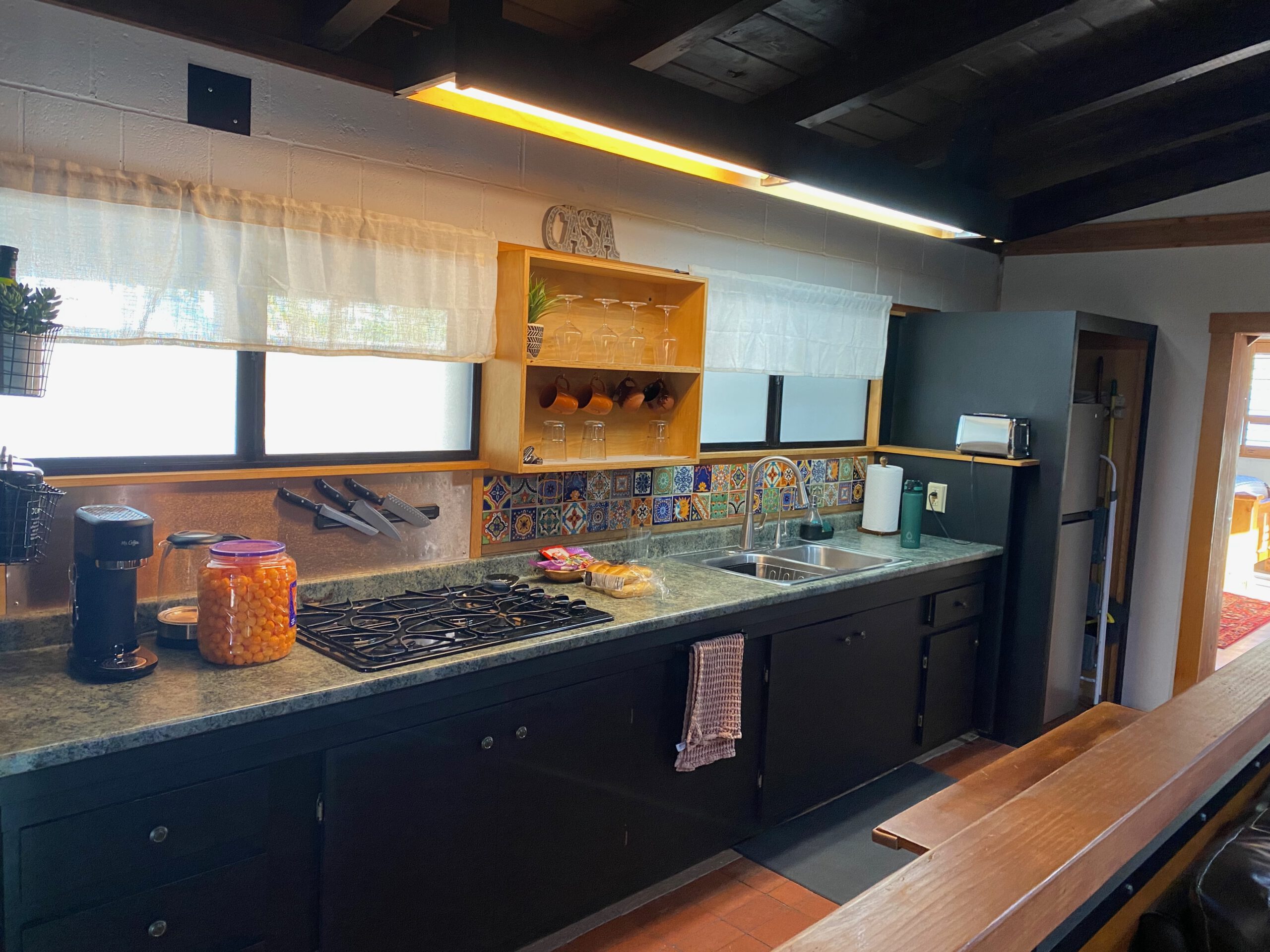 Spacious kitchen at Casa Rustica Morongo Valley Airbnb featuring dark cabinets, gas stove, colorful tile backsplash, double sink, open shelving, and modern yet practical design.