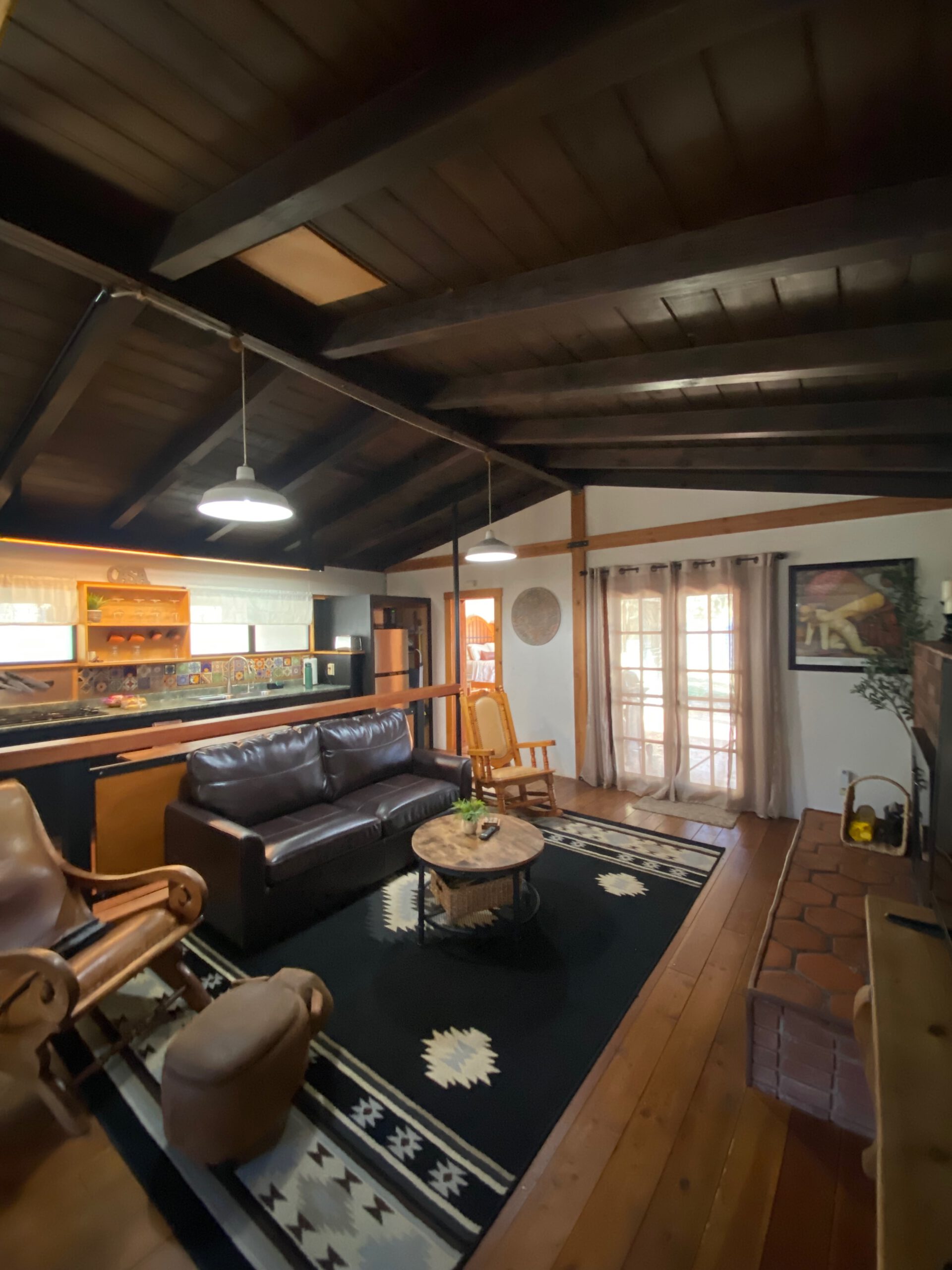 Spacious living room at Casa Rustica Morongo Valley Airbnb with leather sofa, patterned rug, wooden floors, exposed beams, fireplace, and open view toward the kitchen and patio doors.
