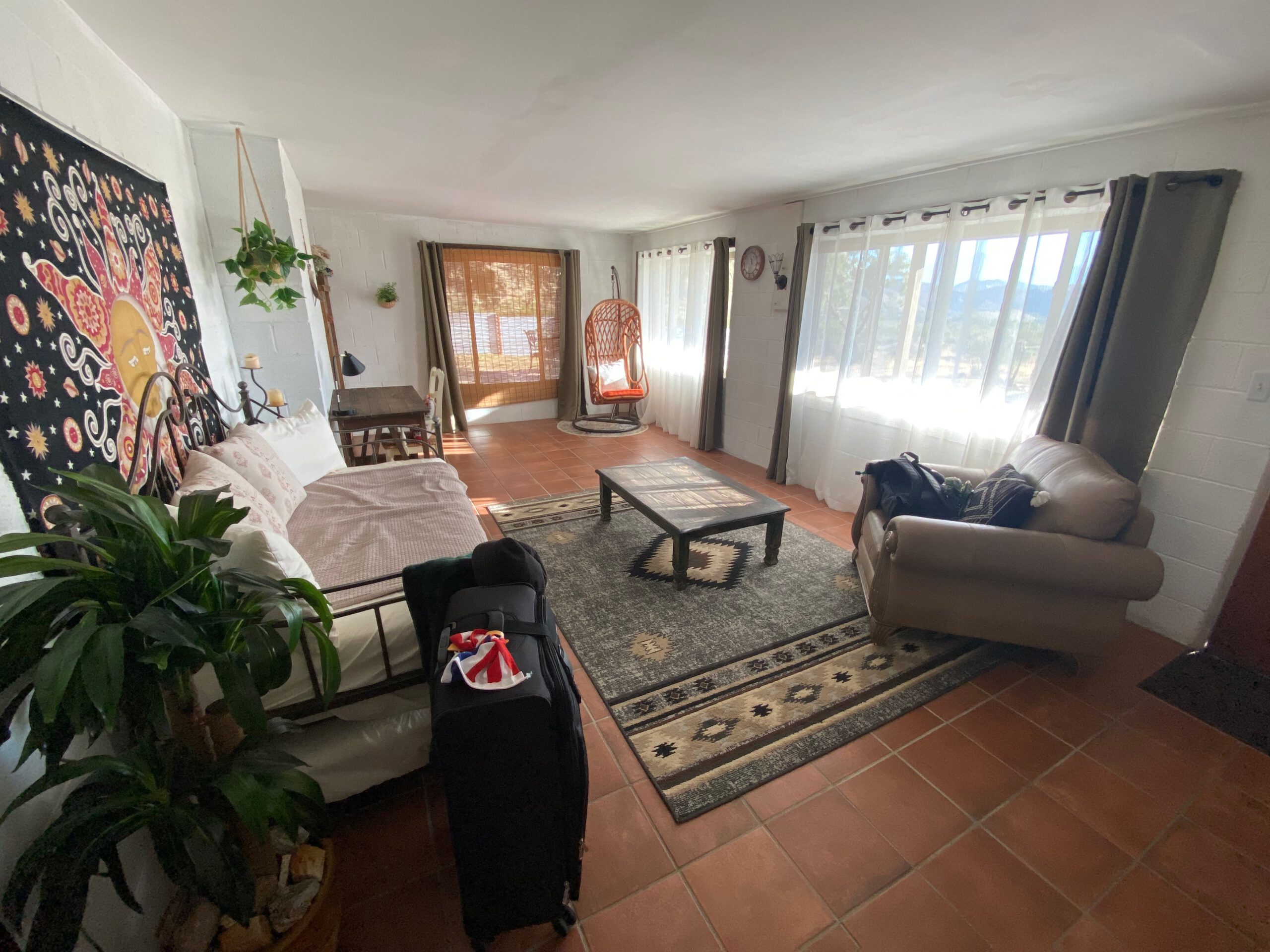 Bright and cozy living room at Casa Rustica Morongo Valley Airbnb with tiled floors, patterned rug, leather sofa, coffee table, hanging chair, natural light, and desert-inspired decor.