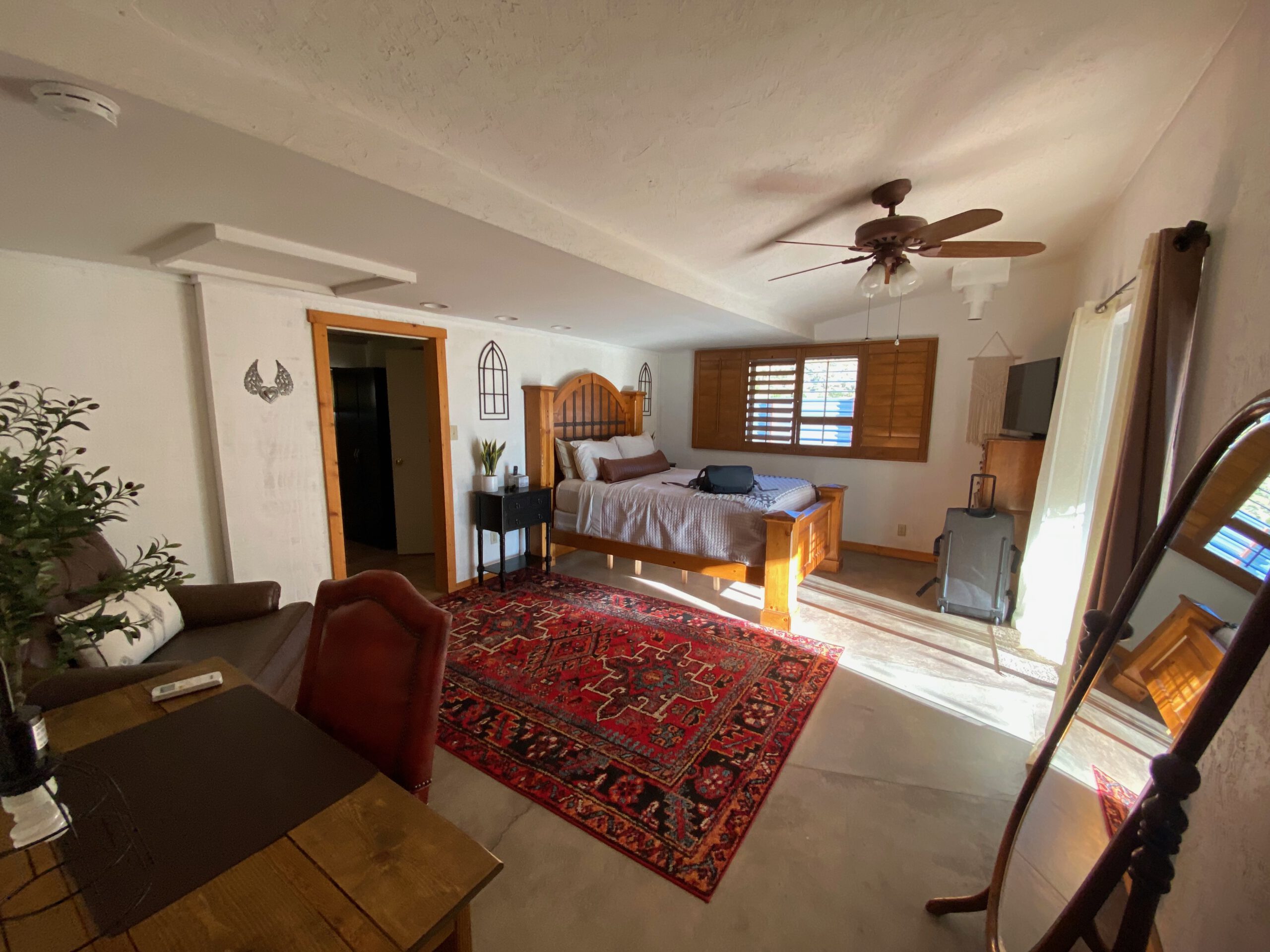 Large main bedroom at Casa Rustica Morongo Valley Airbnb with wooden bed frame, ceiling fan, red patterned rug, seating area, and warm natural light creating a comfortable desert retreat.