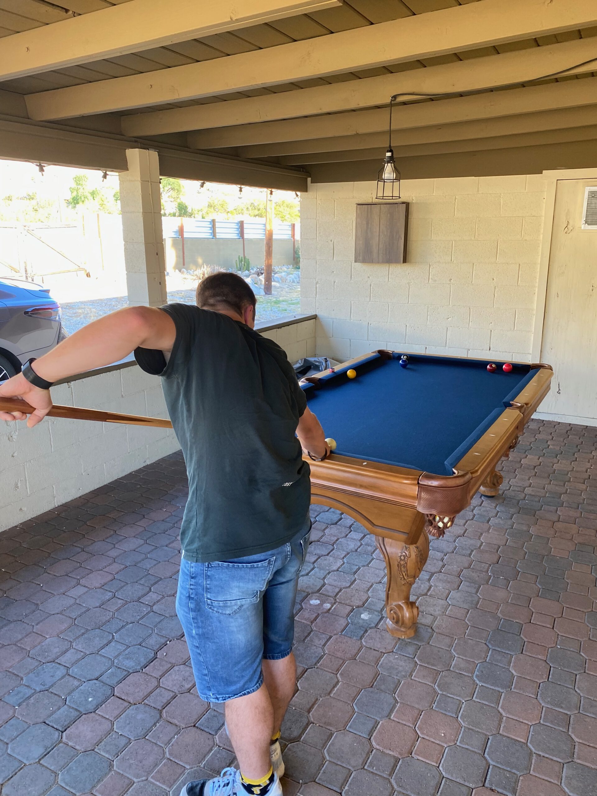 Playing pool at Casa Rustica Morongo Valley Airbnb under a covered outdoor patio, featuring a wooden pool table, brick flooring, and relaxed desert-style setting.
