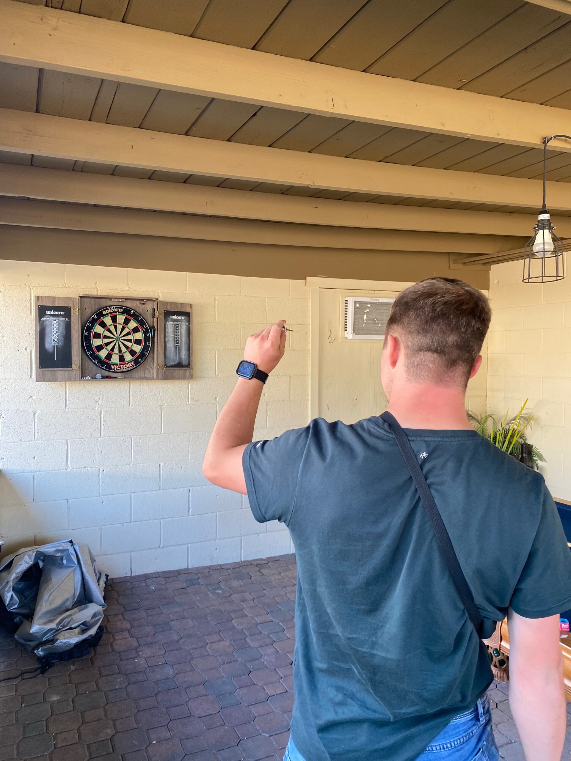 Playing darts at Casa Rustica Morongo Valley Airbnb in a covered outdoor game area with a dartboard mounted on the wall and casual evening atmosphere.
