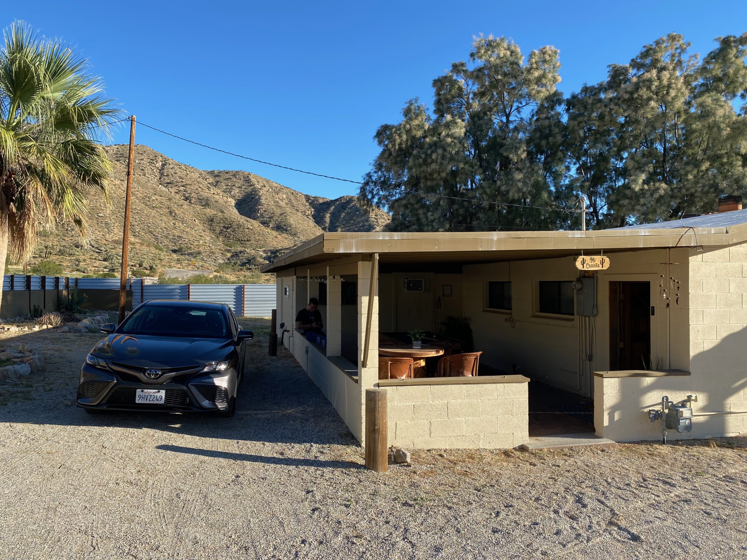 Exterior view of Casa Rustica Morongo Valley Airbnb with private driveway and parked car, covered outdoor seating area, desert-style house, palm tree, and rocky hills in the background under a clear blue sky in Morongo Valley, California.