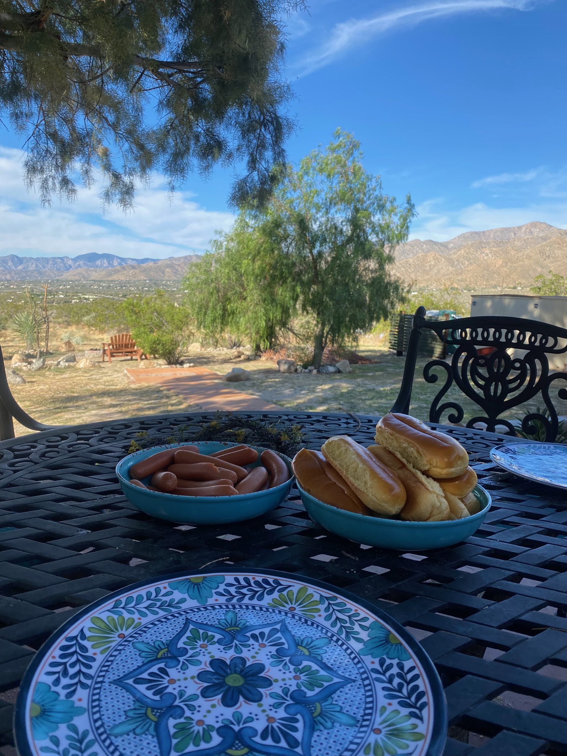 Outdoor dining table at Casa Rustica Morongo Valley Airbnb with hot dog buns and sausages ready for grilling, overlooking a desert garden with trees, mountains, and clear blue sky in Morongo Valley, California.