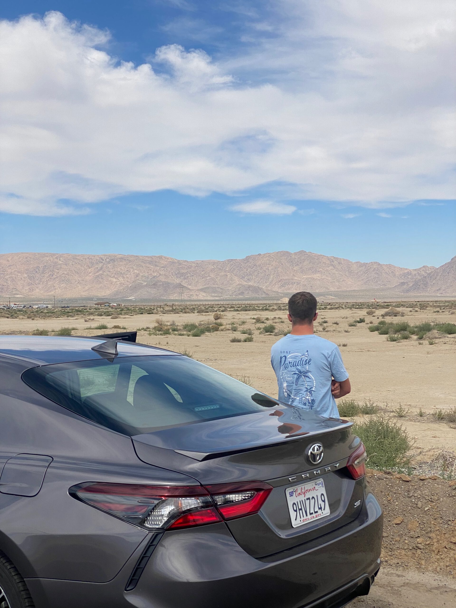 Mojave Desert California road trip stop with a parked car at the roadside, overlooking a wide Mojave Desert landscape with dry plains, scattered shrubs, and distant mountain ranges under a partly cloudy sky.