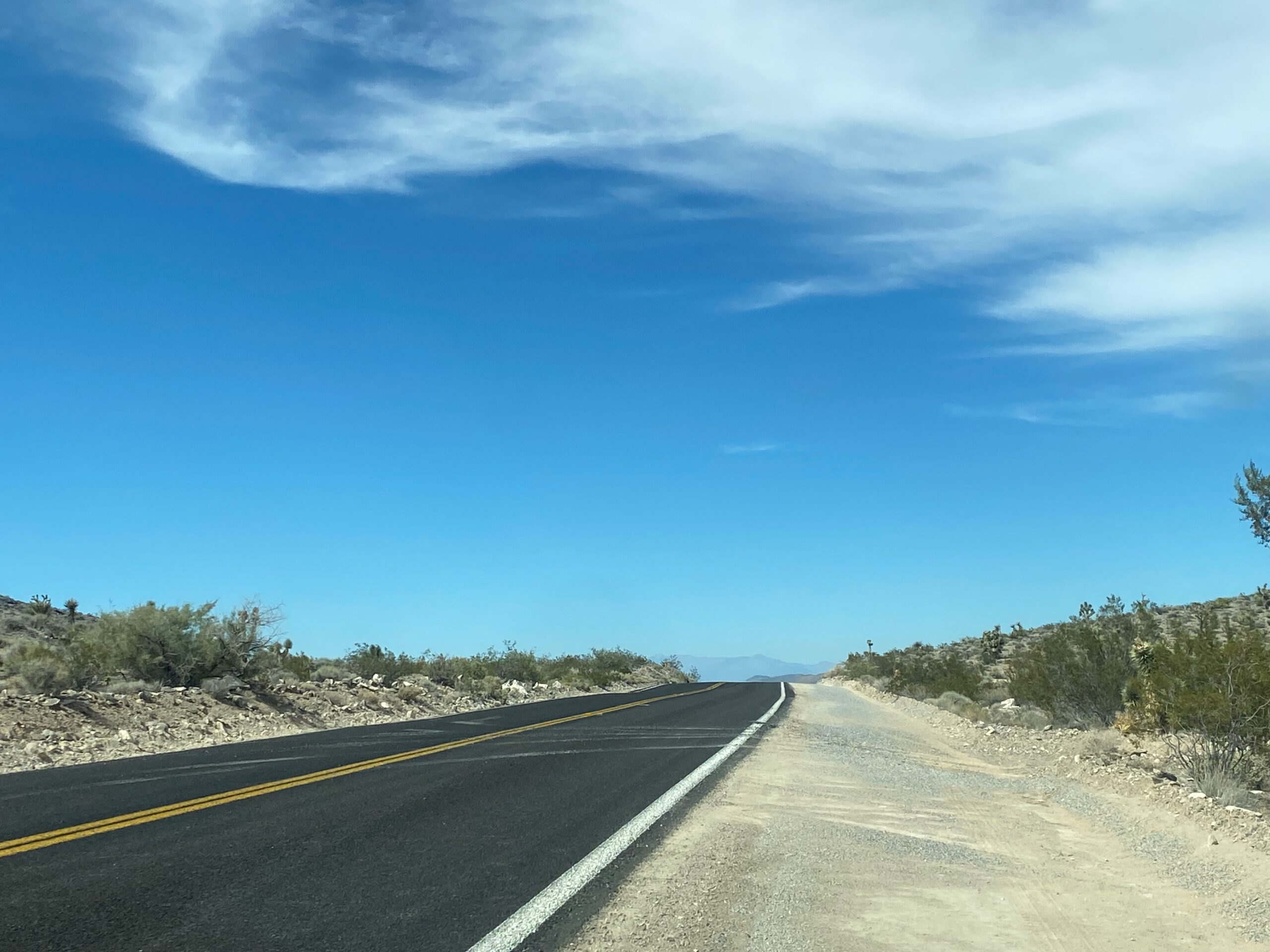 Driving through the Mojave Desert on a quiet paved road, surrounded by dry Mojave Desert landscape, low desert vegetation, and a clear blue California sky.