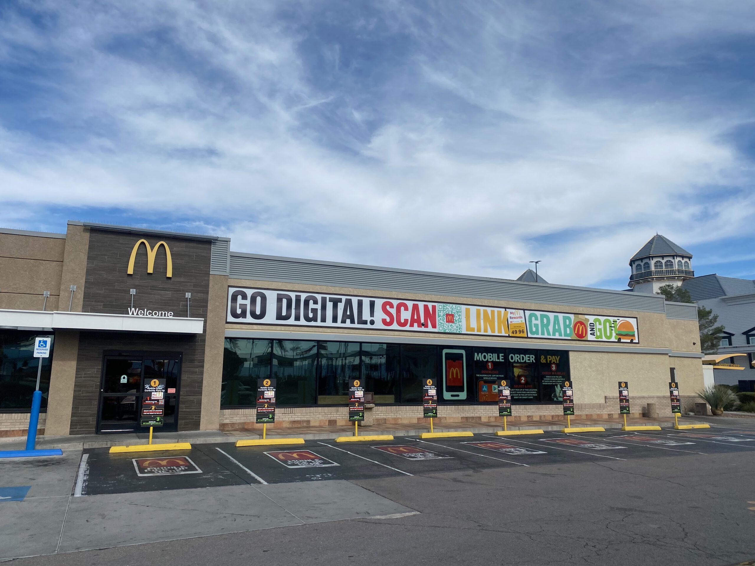 Exterior of McDonald’s Primm Nevada at the California–Nevada border, showing the modern facade with ‘Go Digital’ signage, large parking area, and self-order pickup spots under a partly cloudy desert sky.