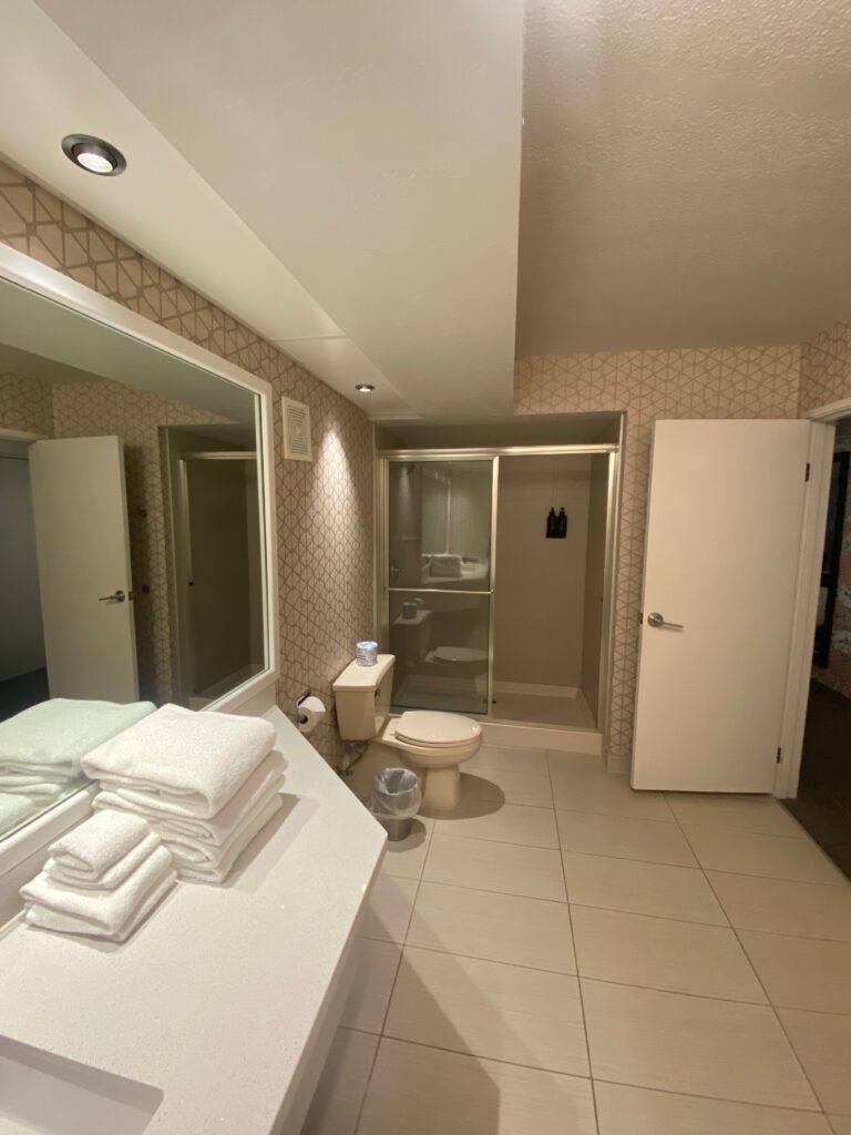 Modern hotel bathroom with a wide vanity, stacked white towels, large mirror, tiled floor, toilet, and a glass-enclosed shower, photographed in clean neutral tones.