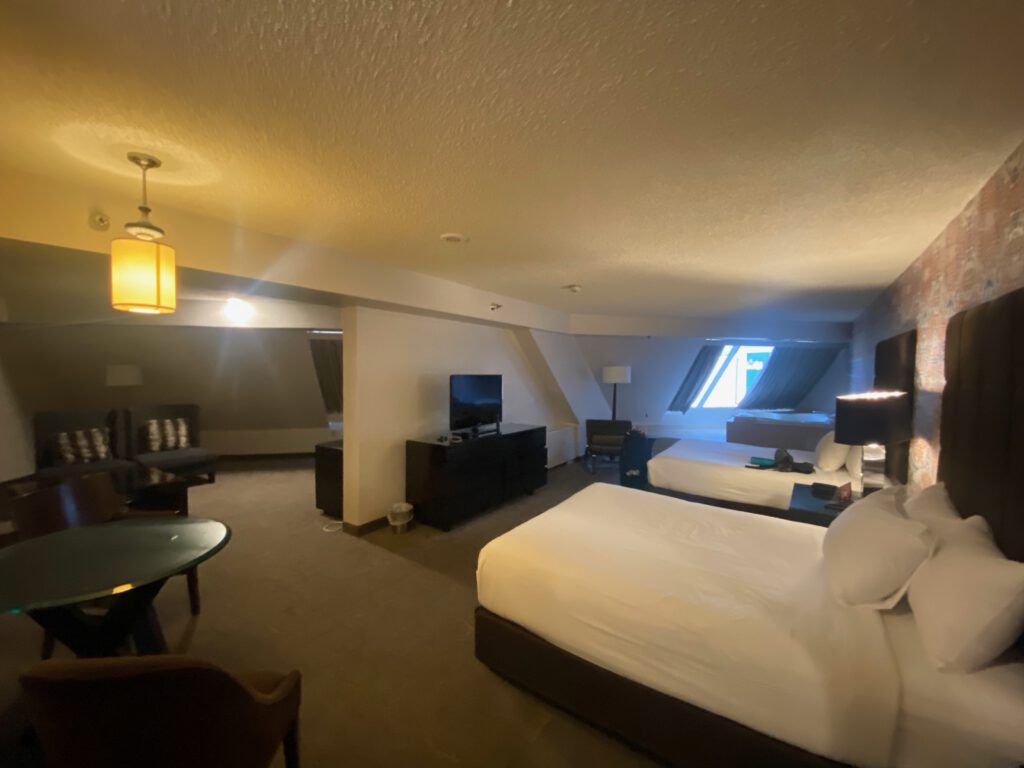 Spacious hotel room with two large beds, seating area, TV cabinet, slanted ceiling architecture, and warm ambient lighting, giving a comfortable Las Vegas suite atmosphere.