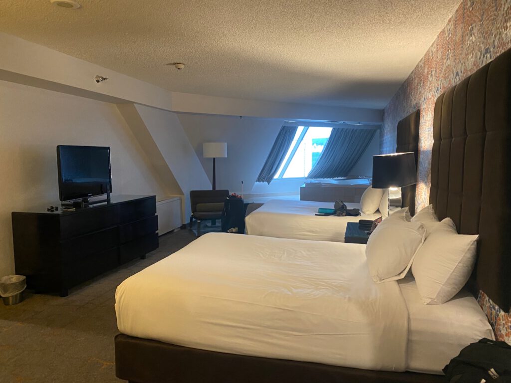 Two neatly made hotel beds facing a slanted window with city views, featuring padded headboards, bedside lamps, and modern Las Vegas hotel room styling.