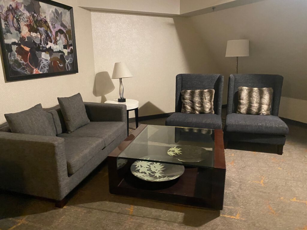 Hotel suite seating area with a gray sofa, two armchairs, glass coffee table, decorative pillows, and abstract wall art, designed for relaxing between city adventures.