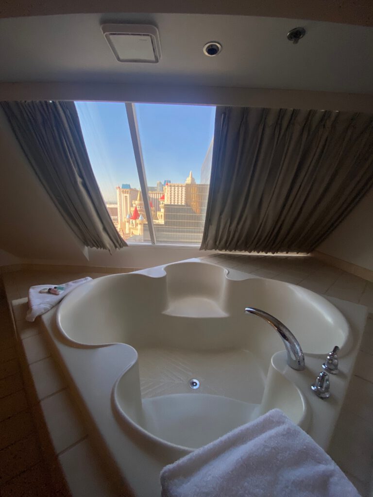 Large sculpted soaking bathtub placed under a slanted window, overlooking the Las Vegas skyline, with soft daylight, towels, and a calm luxury hotel vibe.