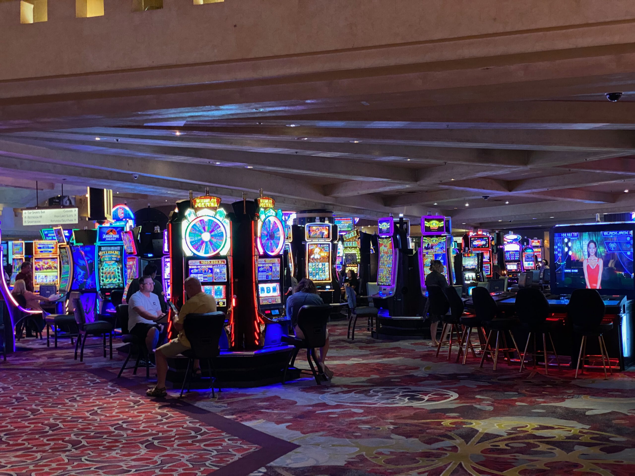 Interior of a Las Vegas casino with rows of brightly lit slot machines, colorful LED displays, patterned carpet, and visitors seated and playing at night.