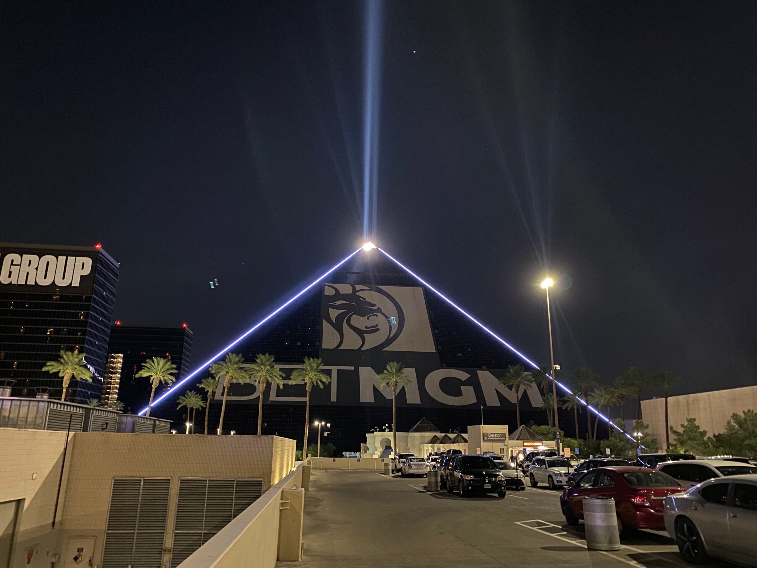 The Luxor pyramid on the Las Vegas Strip at night, featuring the iconic Sky Beam shooting into the dark sky, palm trees in front, and parked cars near the hotel entrance.