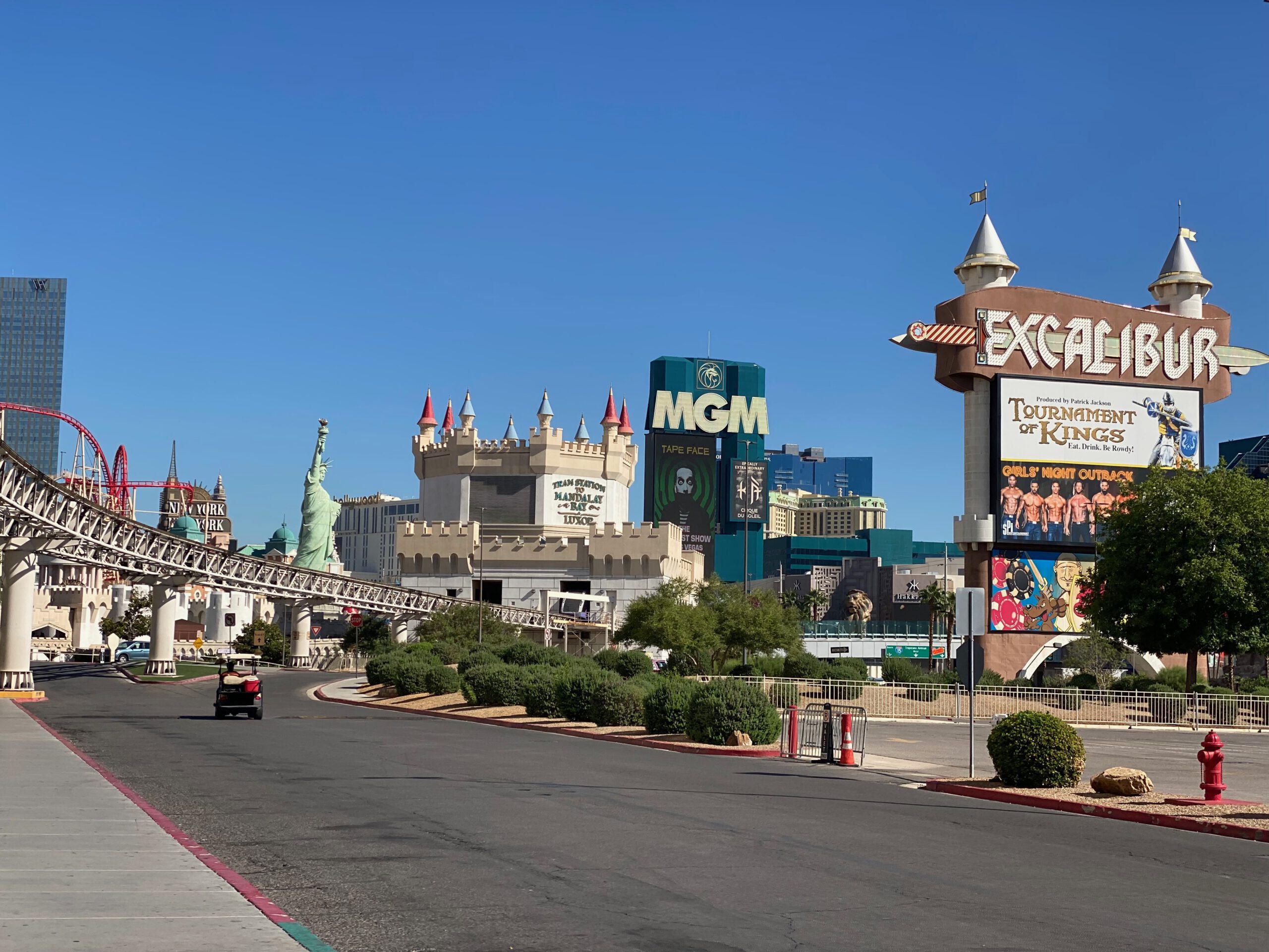 Daytime view of the Las Vegas Strip featuring the Excalibur Hotel & Casino sign, medieval-style towers, the New York-New York roller coaster, the Statue of Liberty replica, and the MGM Grand sign under a clear blue sky.