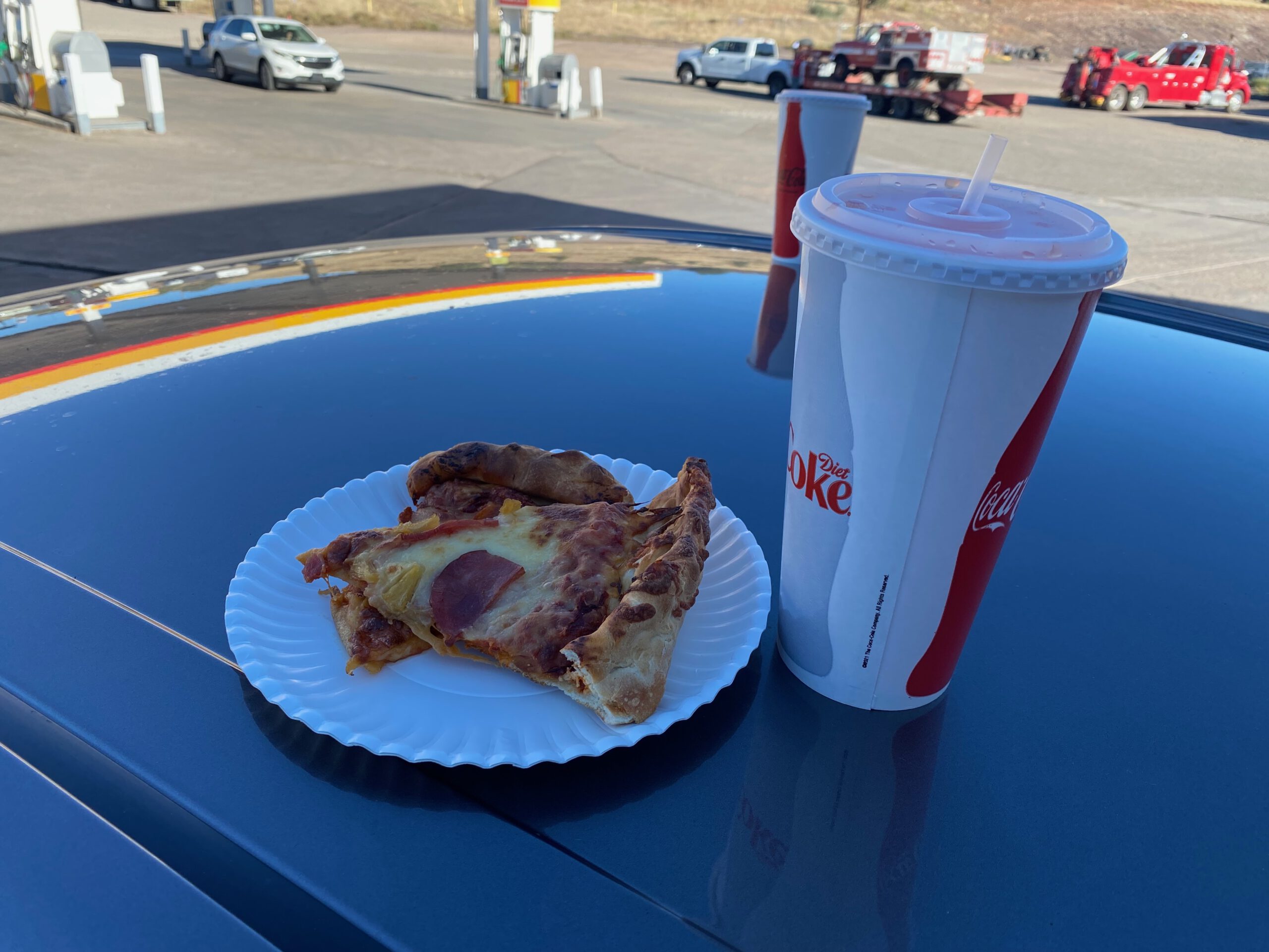 Slice of pizza on a white paper plate next to a large Diet Coke cup, placed on the hood of a car at a Shell gas station in Arizona, with trucks, cars, and fuel pumps visible in the background under clear daylight.