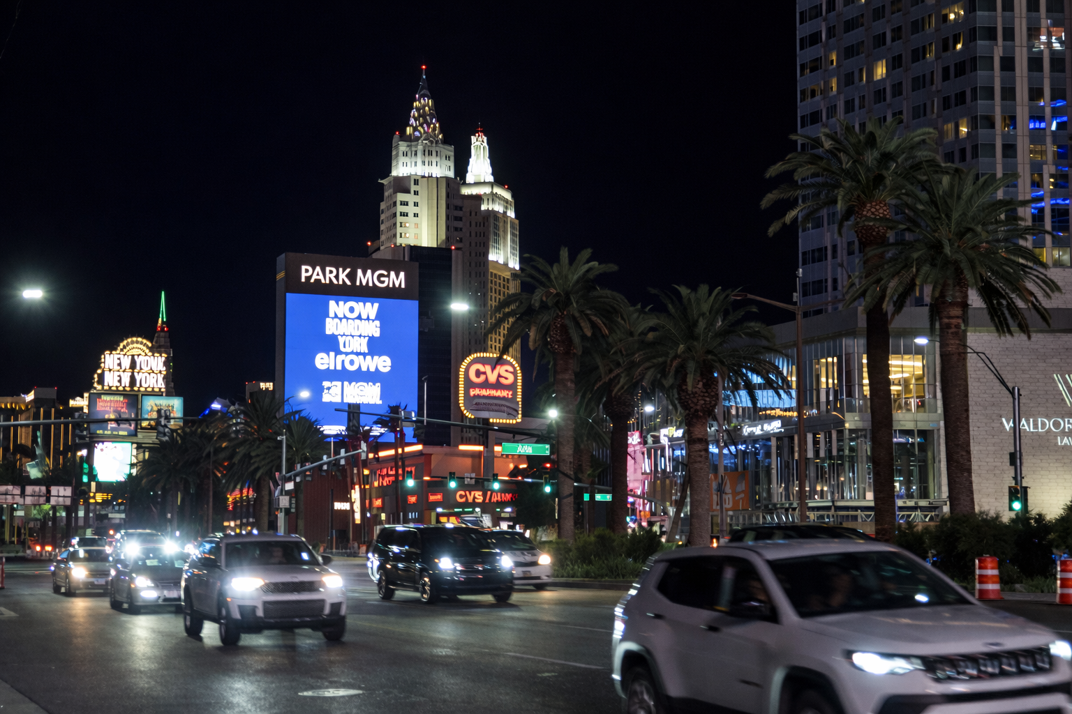 Night view of the Las Vegas Strip with traffic on Las Vegas Boulevard, palm trees lining the street, illuminated signs for Park MGM and CVS Pharmacy, and iconic Strip hotels glowing against the dark sky.