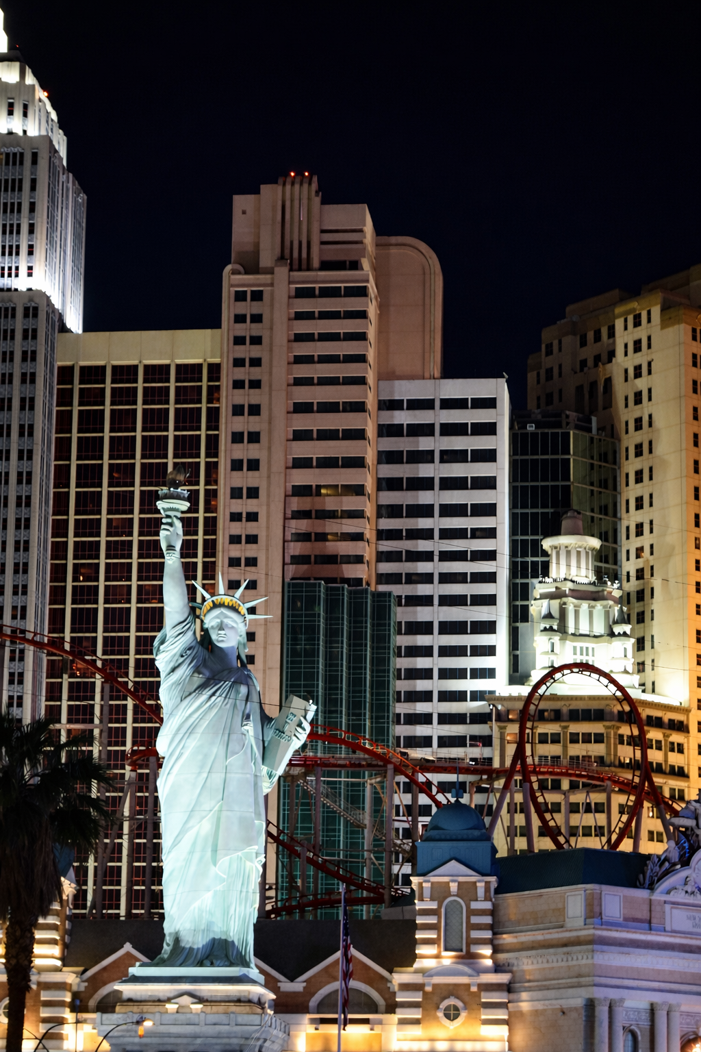 Night view of the Big Apple Coaster at New York-New York Hotel and Casino in Las Vegas, featuring the illuminated Statue of Liberty replica with red roller coaster track looping through a New York–style skyline against a dark sky.