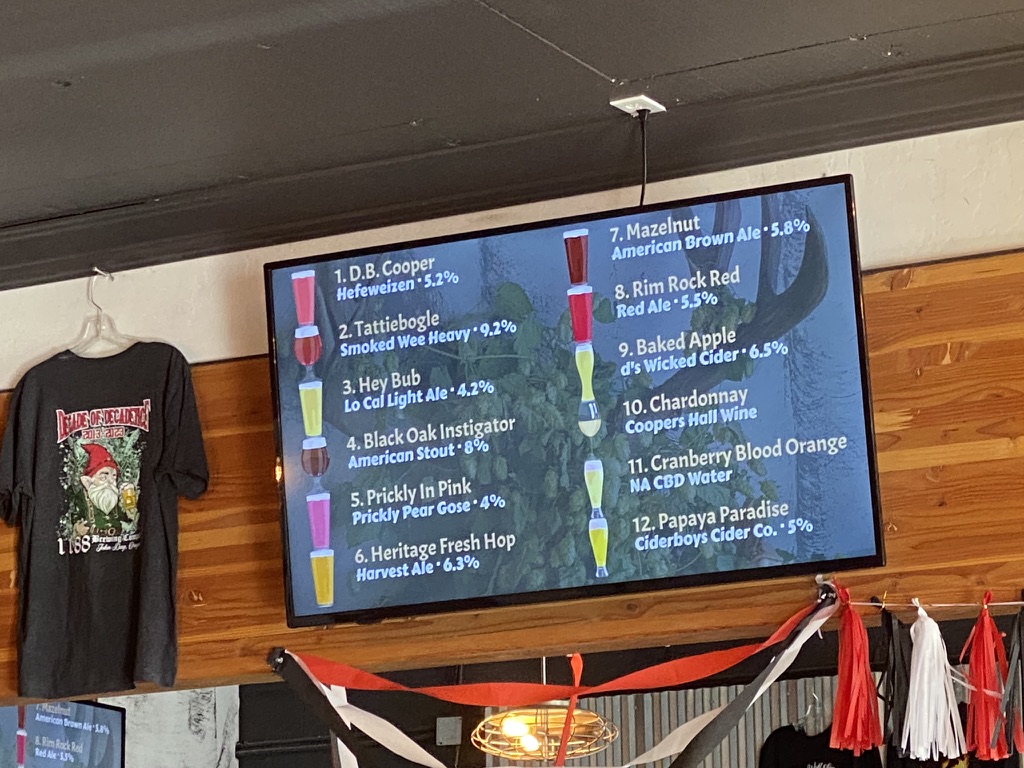 Tap list menu board inside 1188 Brewing Company in John Day Oregon, displaying house brewed beers, seasonal ales, cider options, and wine selections with alcohol percentages in a cozy small town brewery interior.