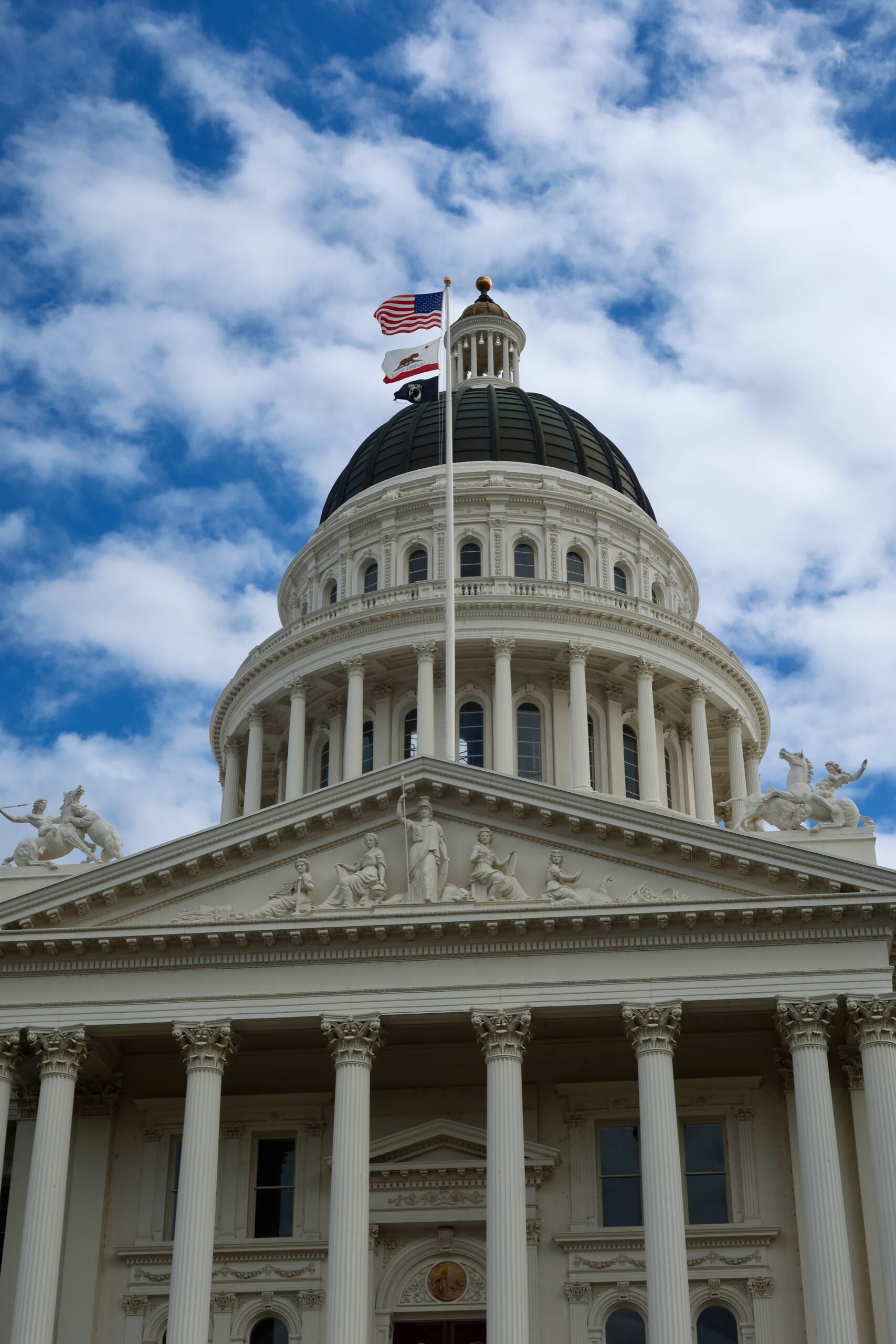 California State Capitol Museum in Sacramento, close up view of the iconic white neoclassical dome with Corinthian columns, sculpted pediment figures, and the United States and California flags against a dramatic blue sky.