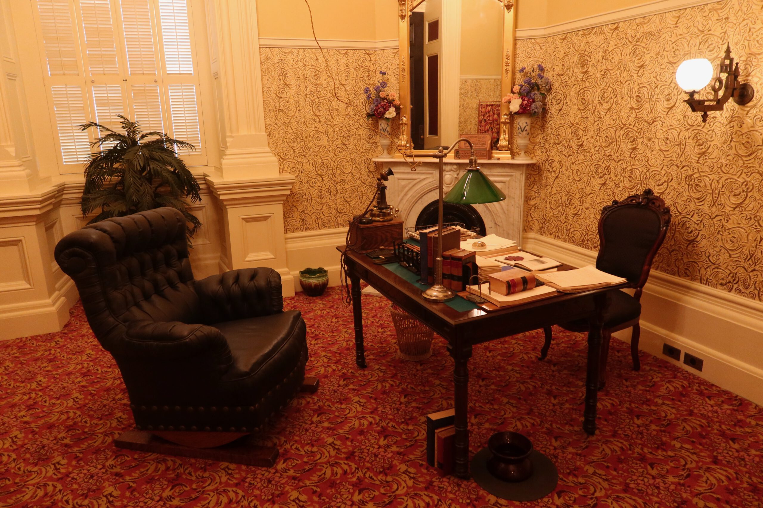 Restored historic office inside the California State Capitol Museum, showing an antique wooden desk, leather armchair, fireplace, books, desk lamp, and patterned wallpaper from nineteenth century California politics.