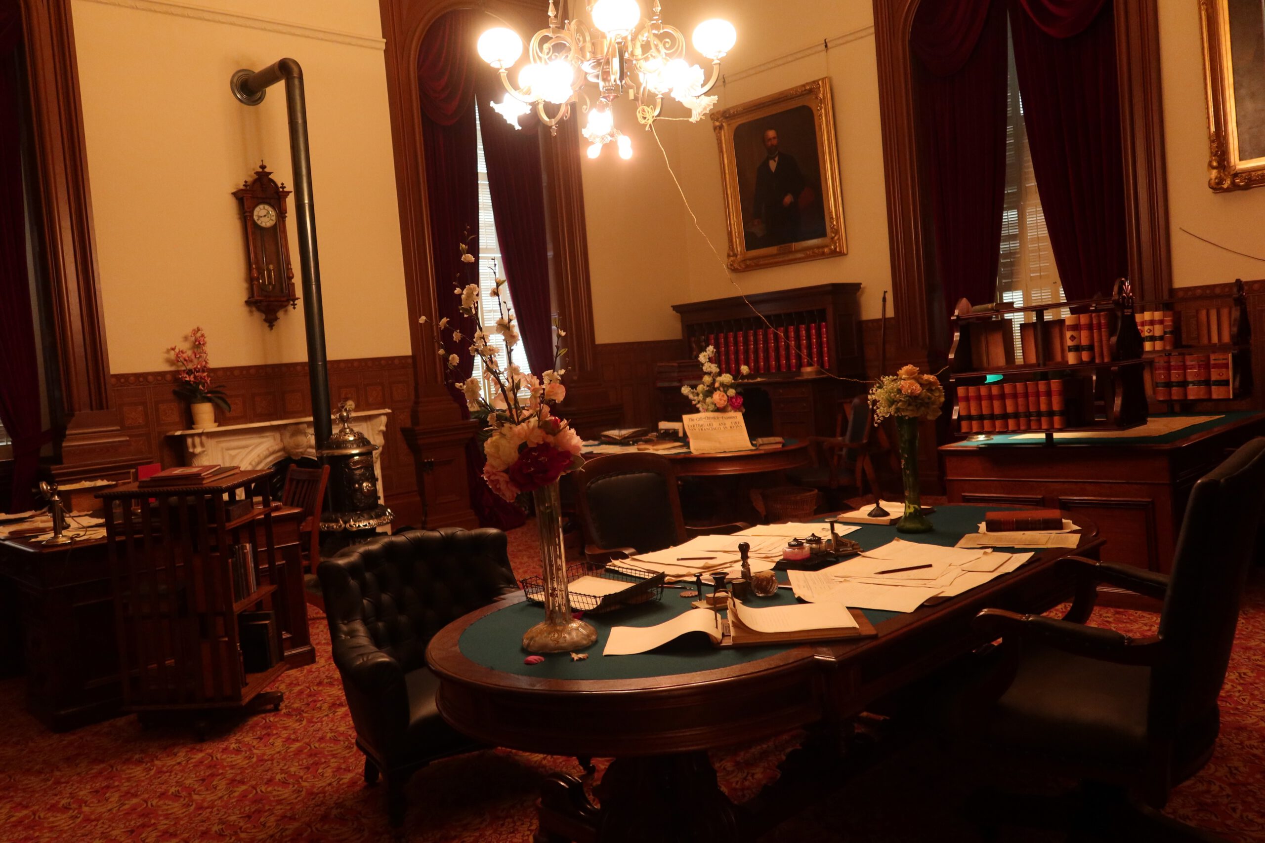 Historic legislative meeting room inside the California State Capitol Museum, featuring dark wood furniture, green felt tables, vintage papers, chandeliers, red drapes, and period decor representing early California government.