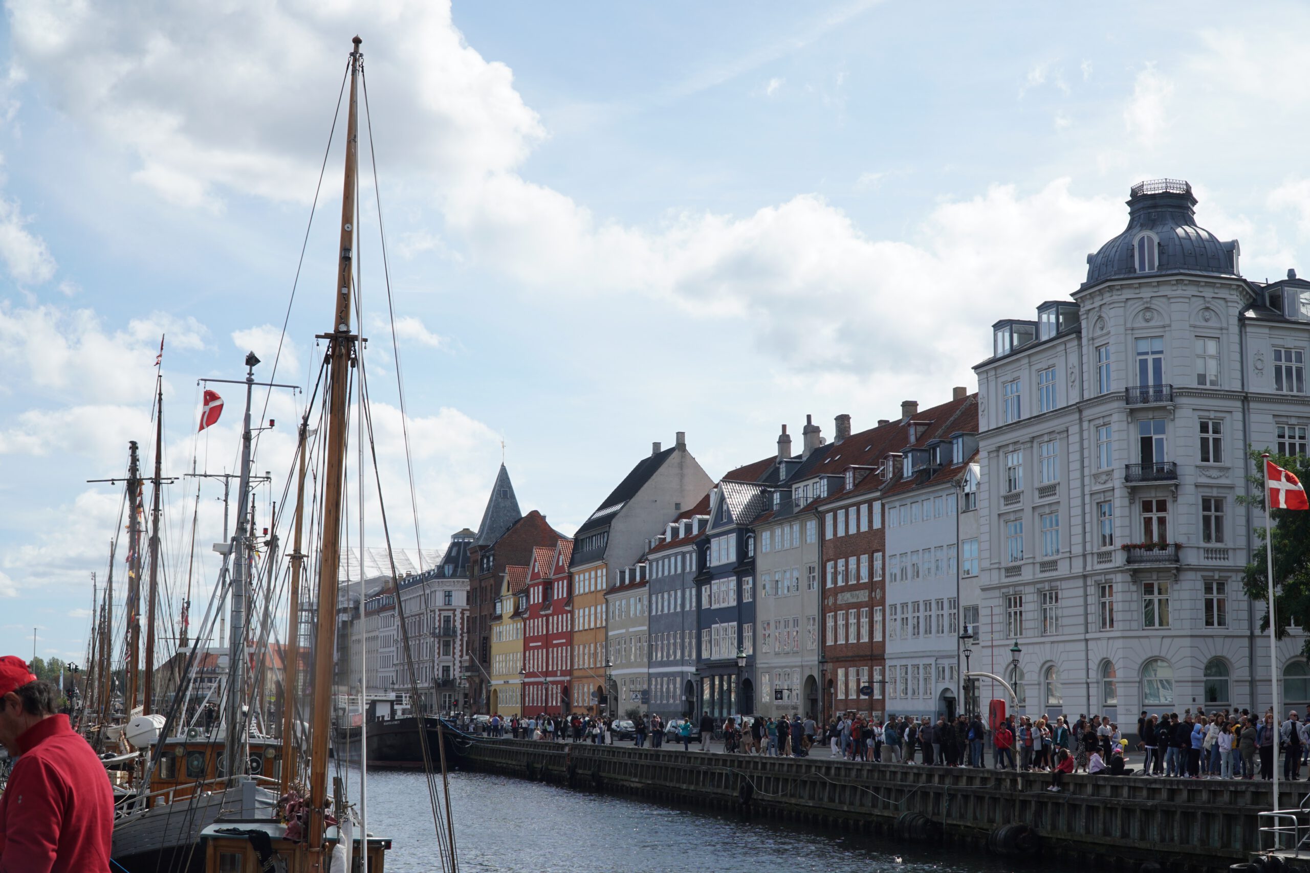 Nyhavn Copenhagen travel guide review, panoramic view of the Nyhavn canal featuring tall masts, classic ships, elegant historic buildings, and a long line of visitors walking along the harbor.