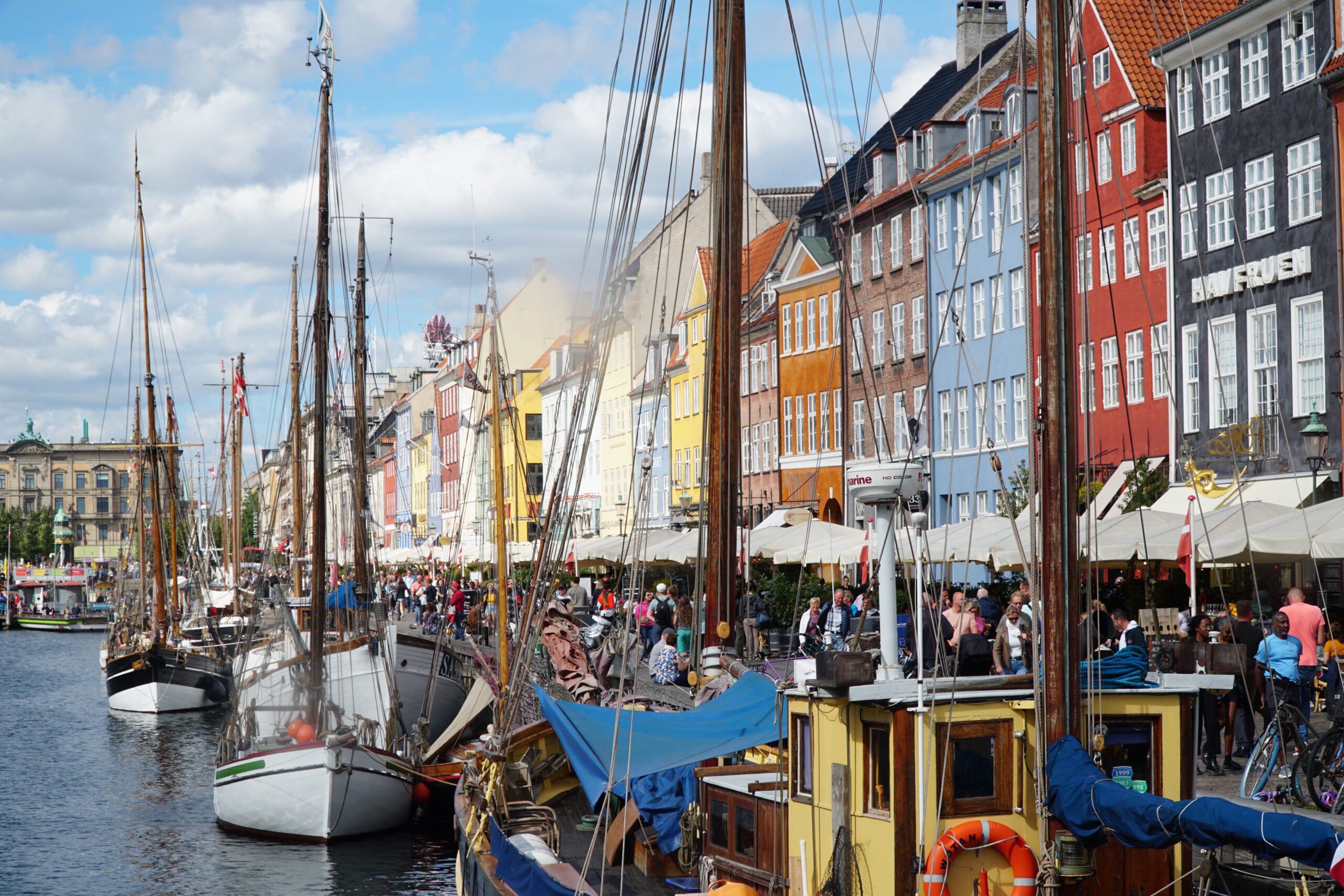 Nyhavn Copenhagen travel guide review, historic wooden ships lined up along the colorful waterfront houses of Nyhavn harbor in Copenhagen on a busy sunny day.