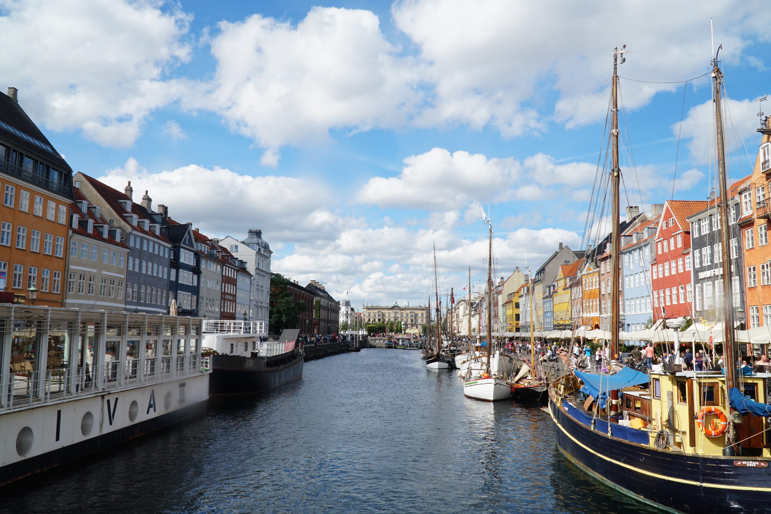 Nyhavn Copenhagen travel guide review, wide view of the Nyhavn canal with historic wooden ships, colorful waterfront houses, busy terraces, and blue skies in central Copenhagen.
