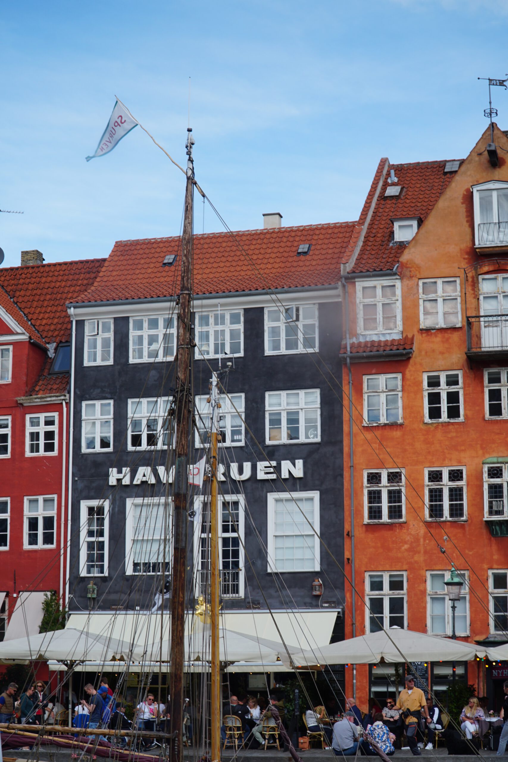 Nyhavn Copenhagen travel guide review, close-up of colorful historic townhouses with red tiled roofs, café terraces, and sailing ship masts along the Nyhavn waterfront in Copenhagen.