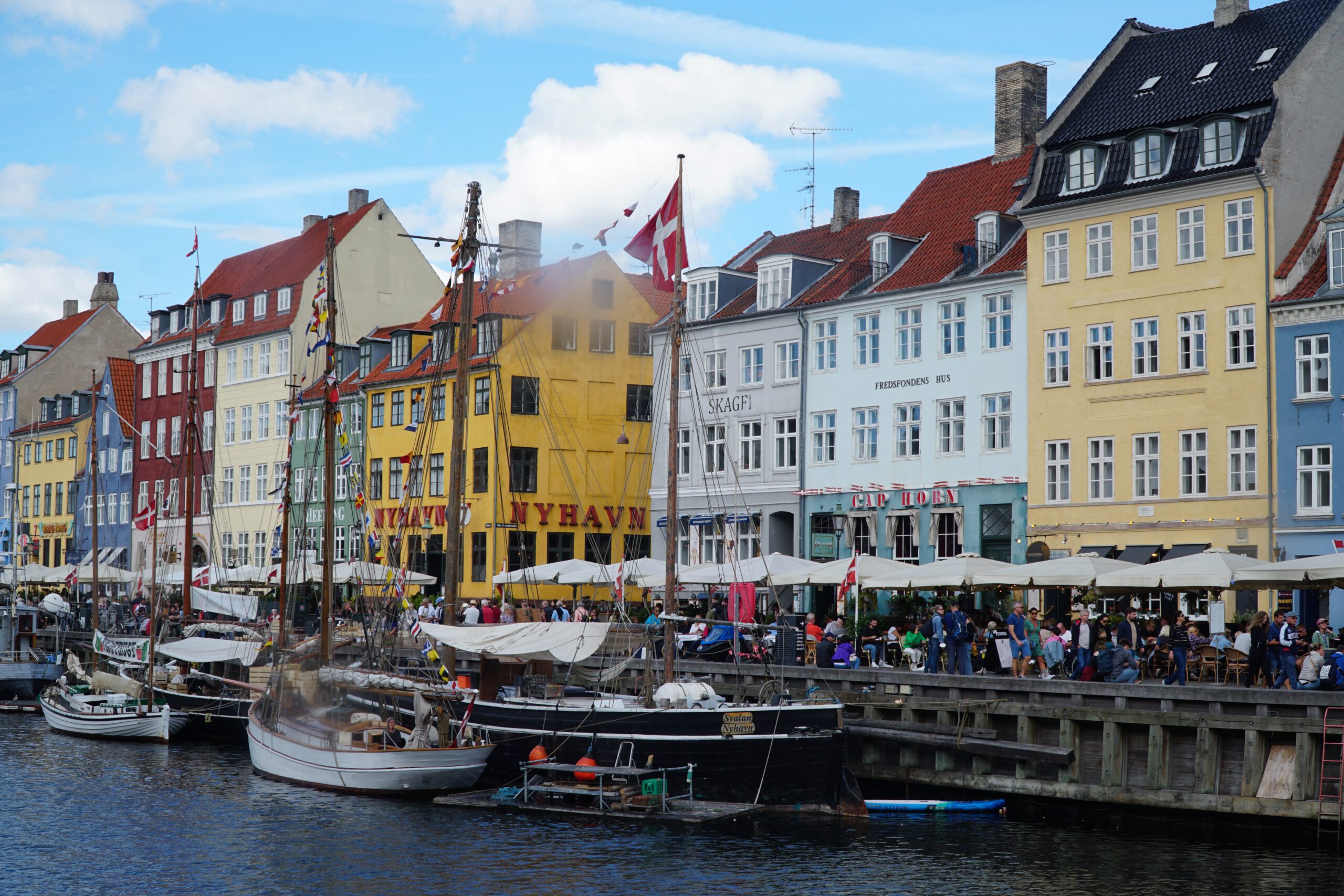 Nyhavn Copenhagen travel guide review, lively waterfront scene with historic sailing boats, colorful 17th-century houses, busy restaurant terraces, and crowds along the canal in Copenhagen.