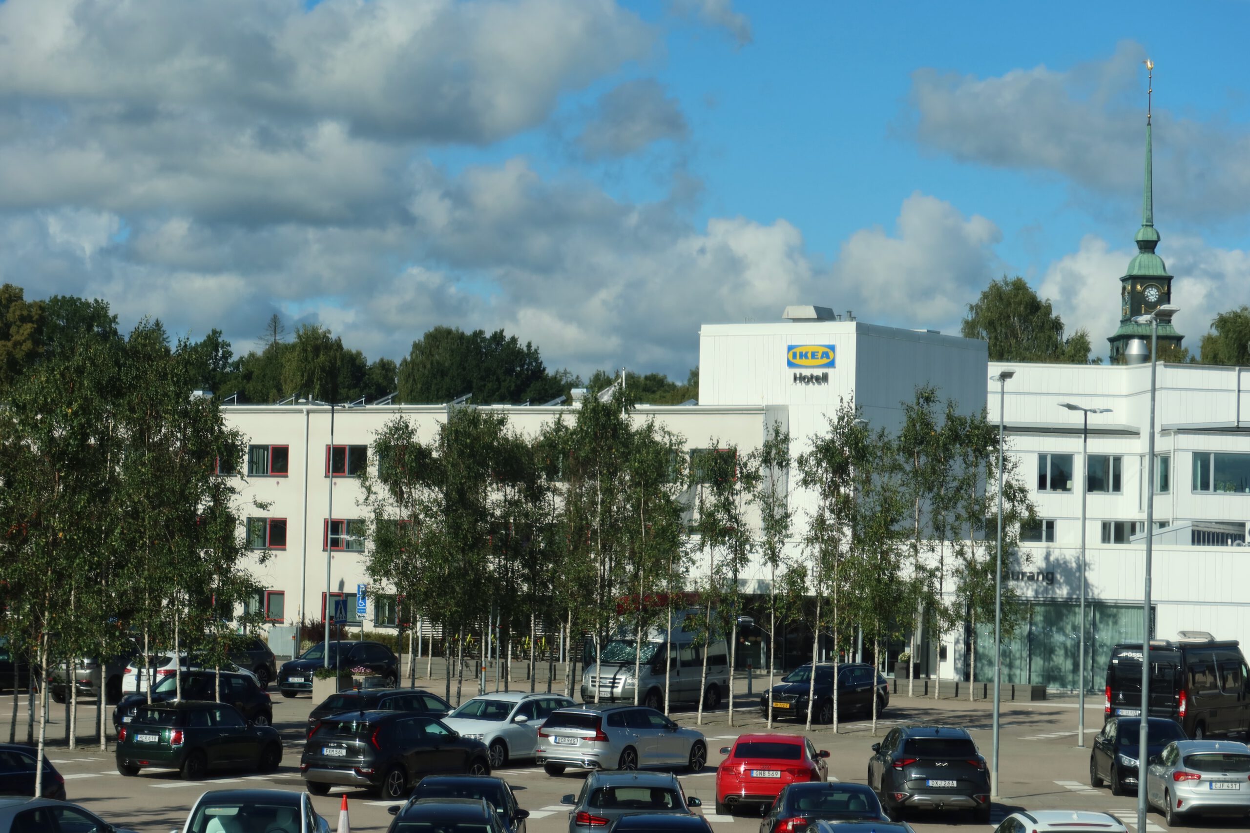 Parking area at IKEA Hotell Älmhult with multiple parked cars, tree-lined spaces and a clear view of the hotel building under a bright blue sky in Älmhult, Sweden.