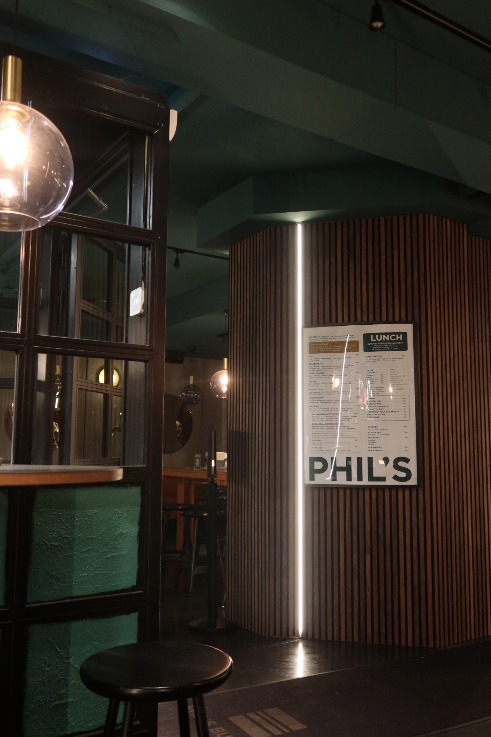 Interior view of Phils Burger Drottninggatan Stockholm, featuring the modern design with wood panel walls, green ceiling, menu display, bar seating, and minimalist lighting inside the restaurant