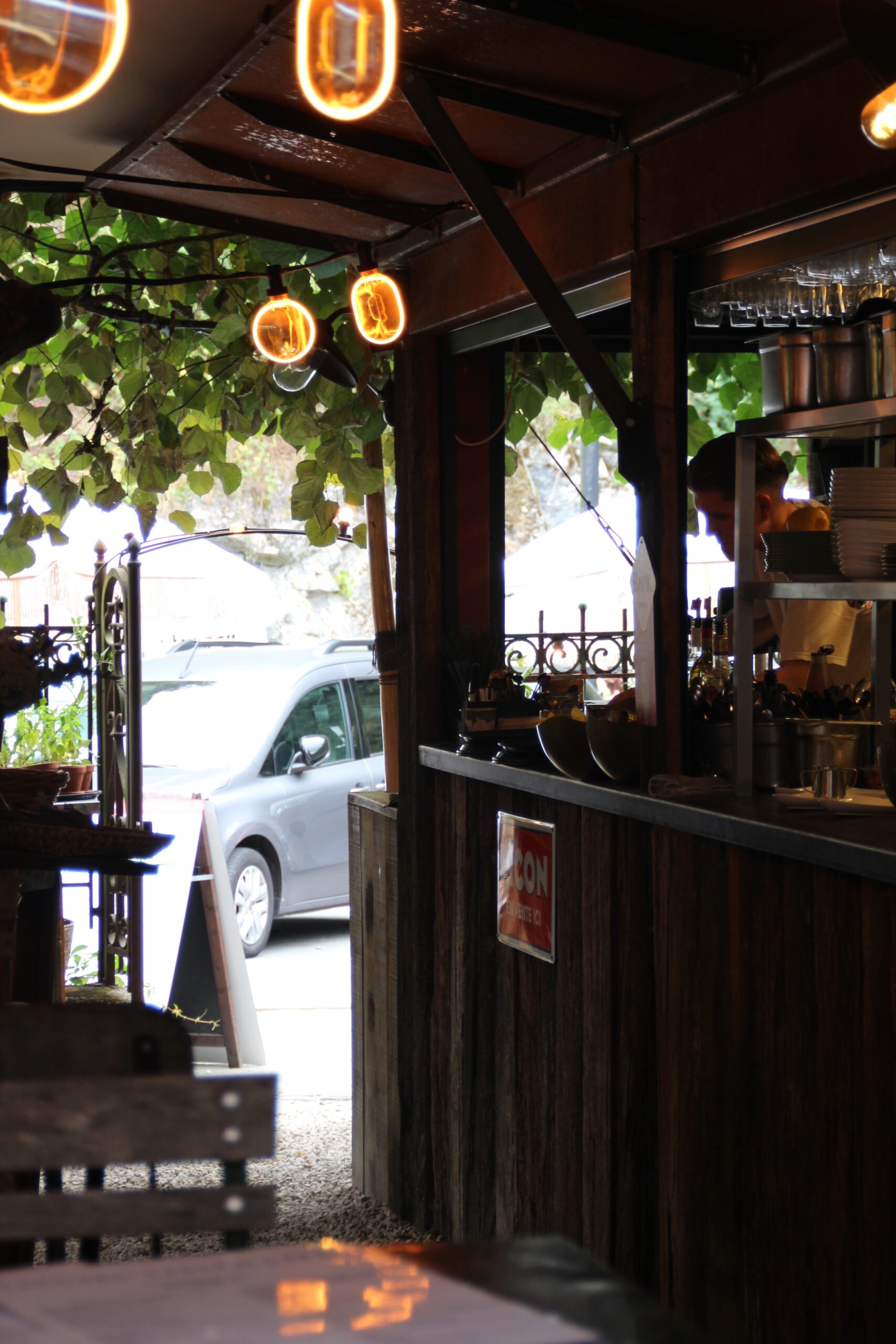Edouard Garden Café Durbuy garden terrace with wooden bar, warm hanging lights, leafy surroundings, and relaxed outdoor dining atmosphere in the center of Durbuy