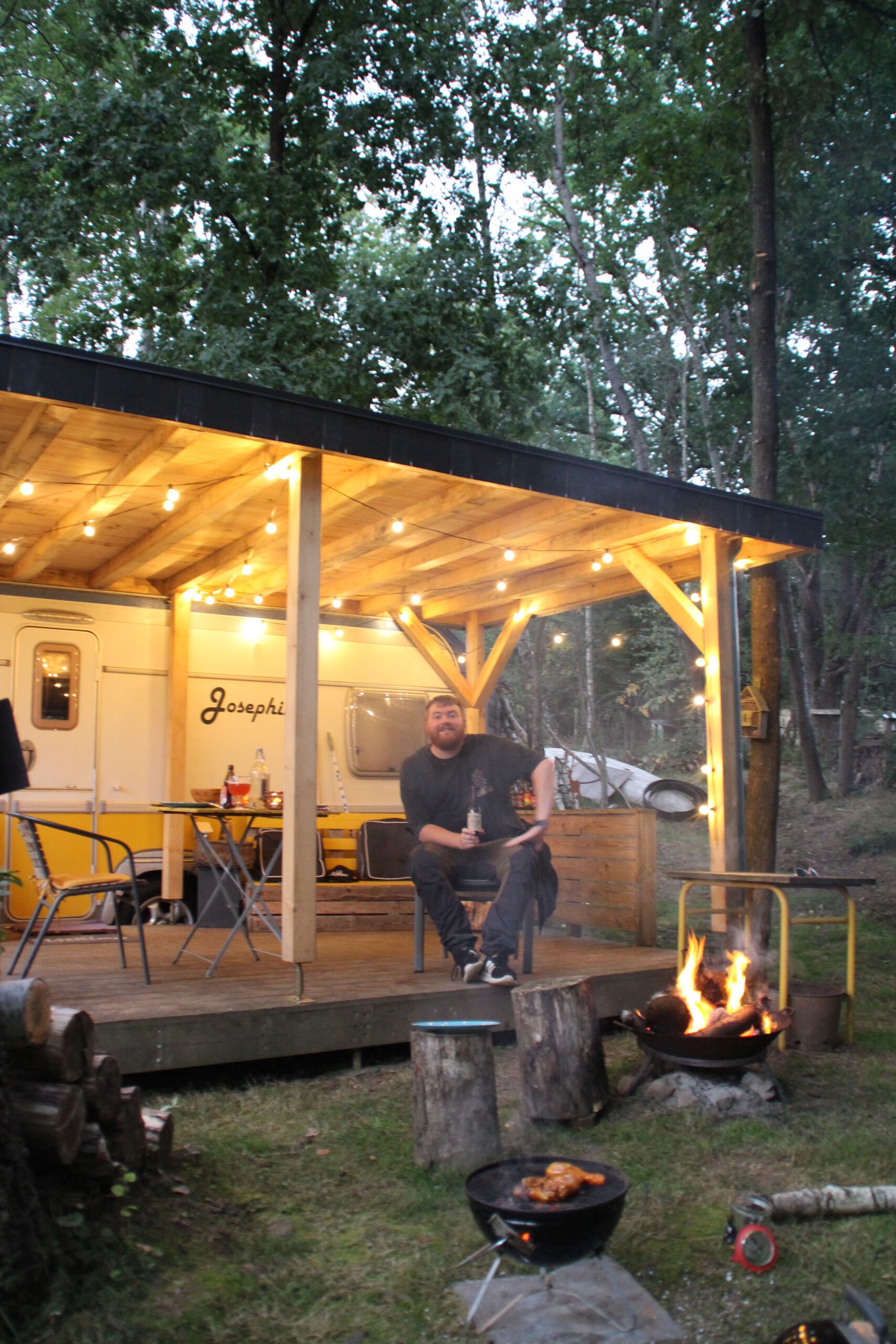 Josephine Caravan Aywaille Timo van den Berg relaxing on the wooden porch beside the fire pit during a cozy evening in the Ardennes