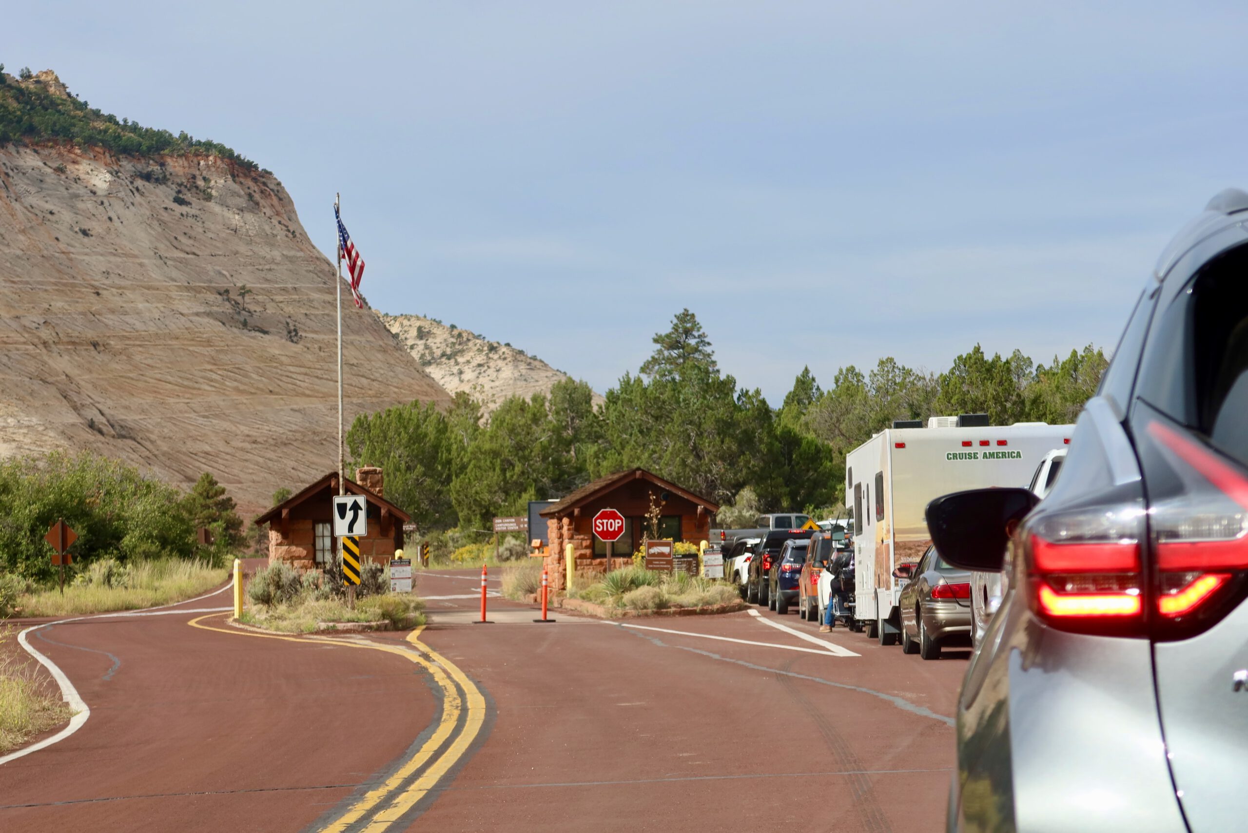 Entrance checkpoint of Zion National Park with cars lined up, ranger station cabins, American flag, and red sandstone cliffs in southern Utah