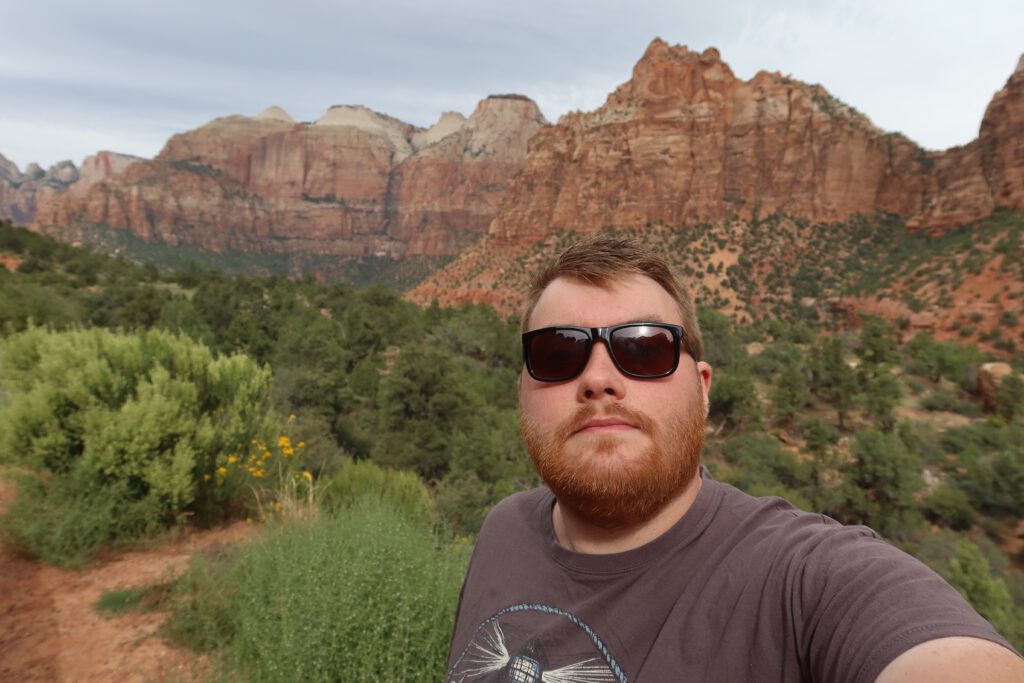 Timo van den Berg (Timeuw) taking a selfie with red sandstone cliffs and green vegetation in Zion National Park, Utah, dramatic canyon landscape in the American Southwest