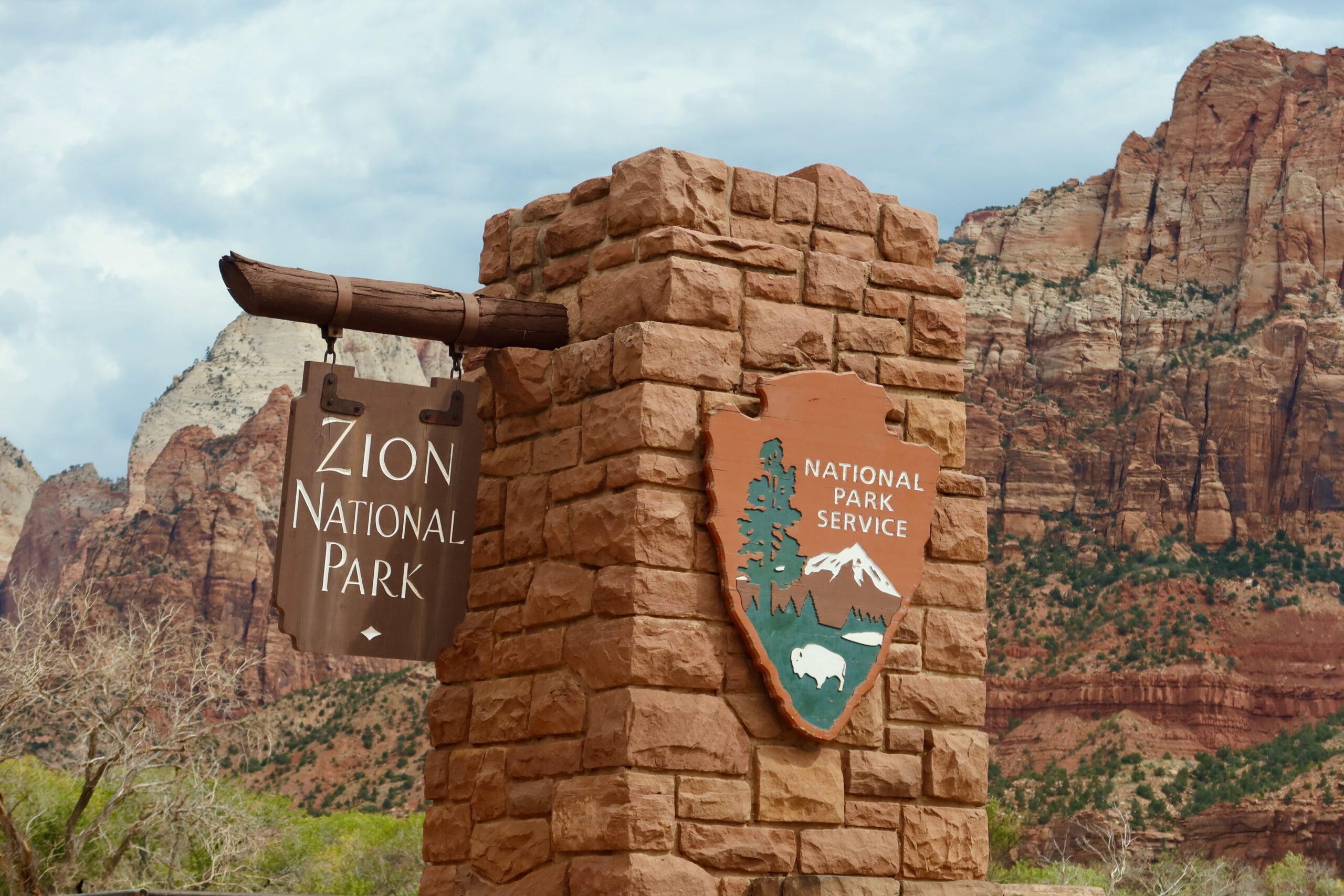 Zion National Park entrance sign with National Park Service emblem, framed by towering red sandstone cliffs in southern Utah