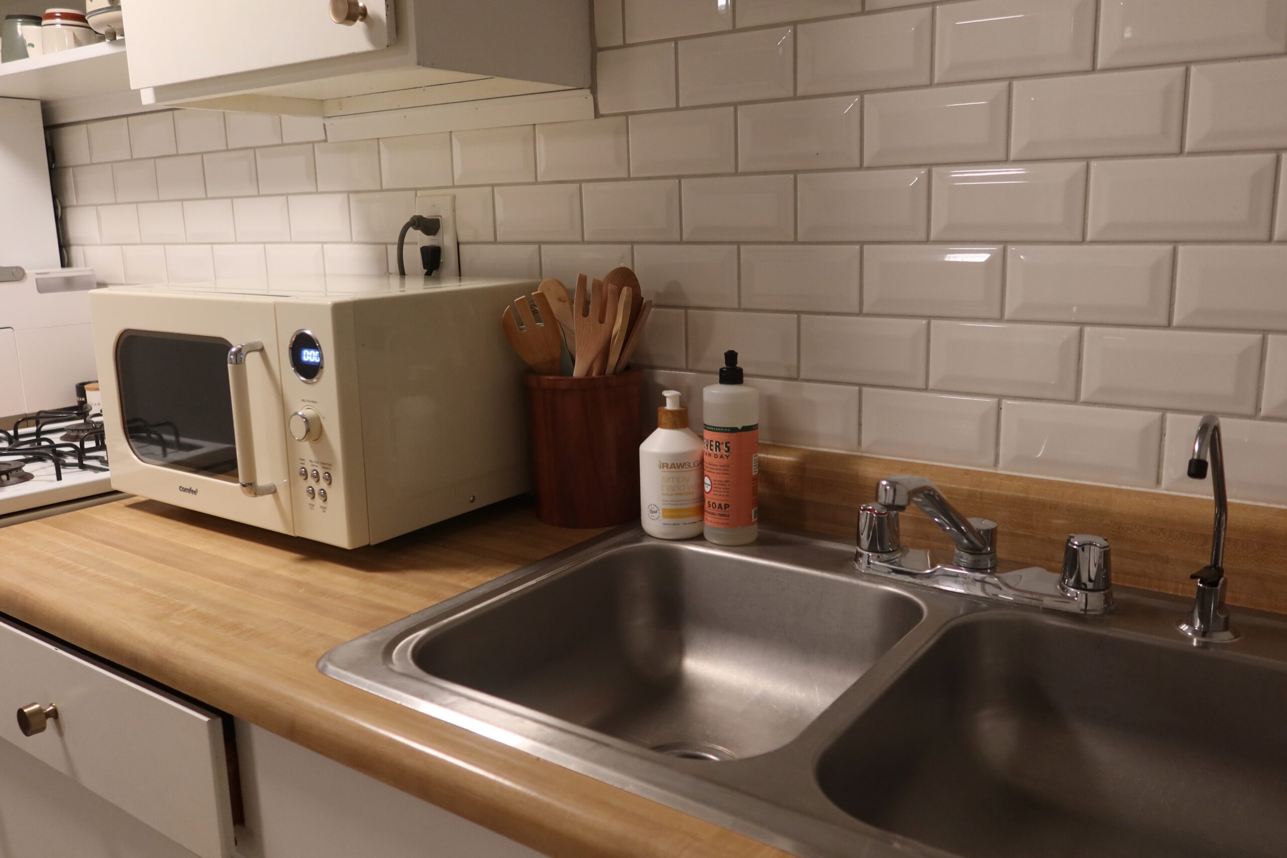 Clean kitchen area in a Sandy Utah Airbnb basement stay, featuring a double sink, gas stove, microwave, wooden countertop, cooking utensils, and soap essentials, ideal for a comfortable Utah road trip stop
