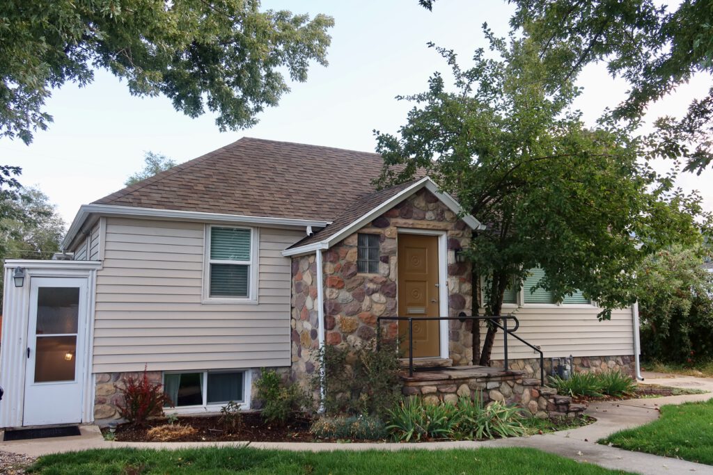 Exterior daytime view of a Sandy Utah Airbnb, showing a quiet residential home with stone facade, private basement entrance, green lawn, and trees, ideal for a peaceful Utah road trip stay