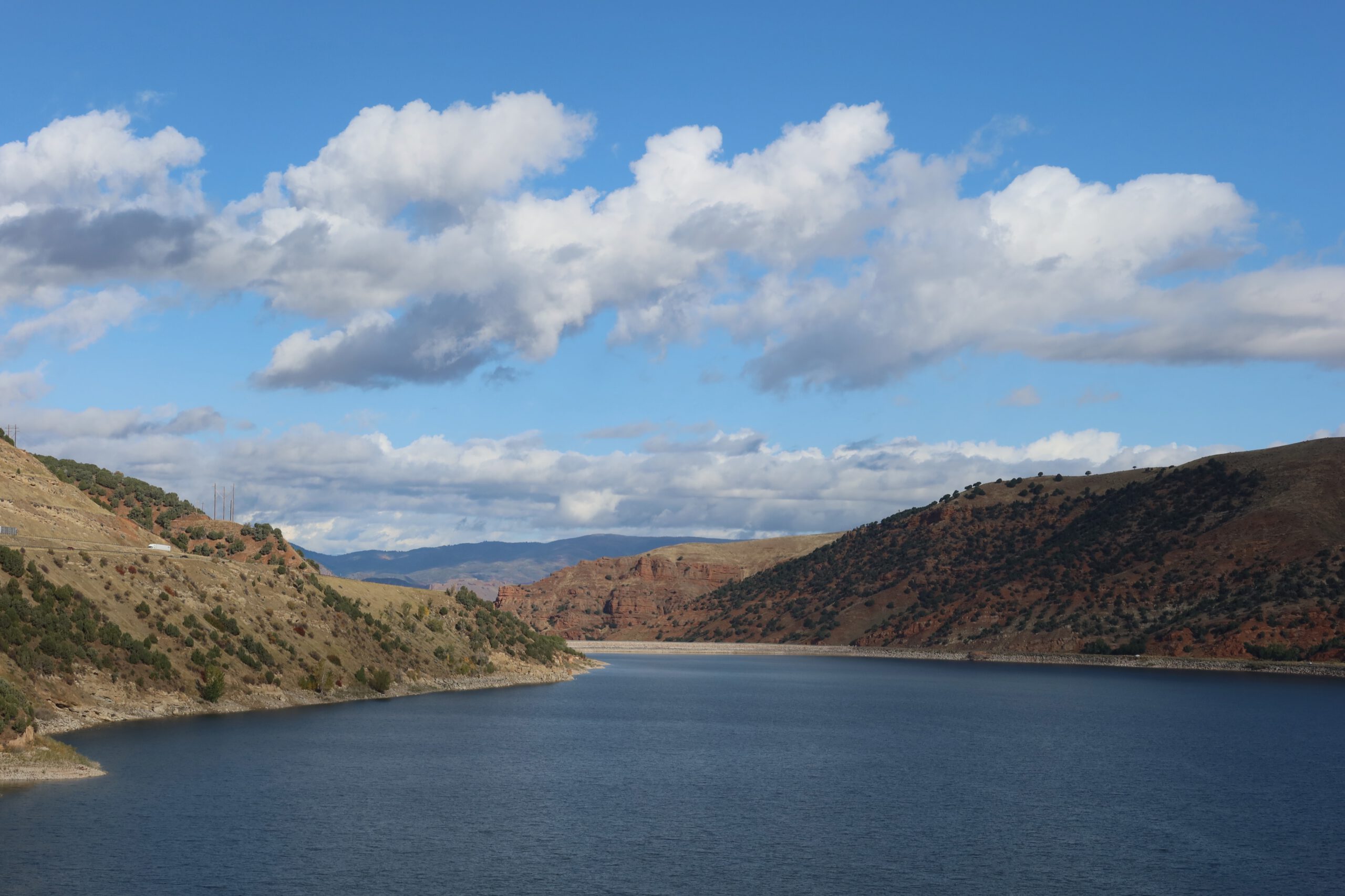 Wide view over Echo Reservoir near Coalville Utah, calm blue water between rugged hills and red rock formations under drifting white clouds.