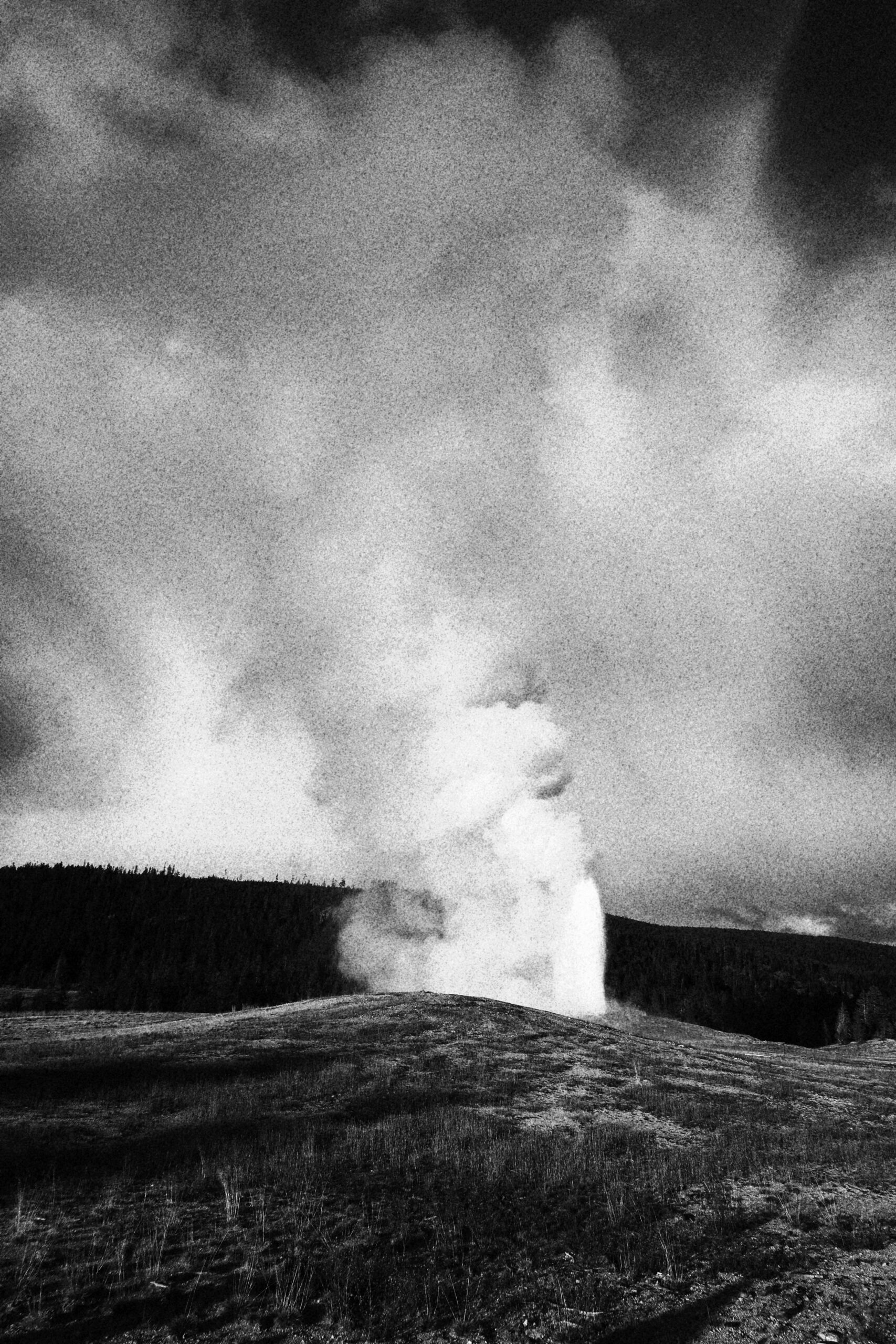 Black and white photo of a geyser erupting in Yellowstone National Park, dramatic steam plume rising under dark cloudy sky