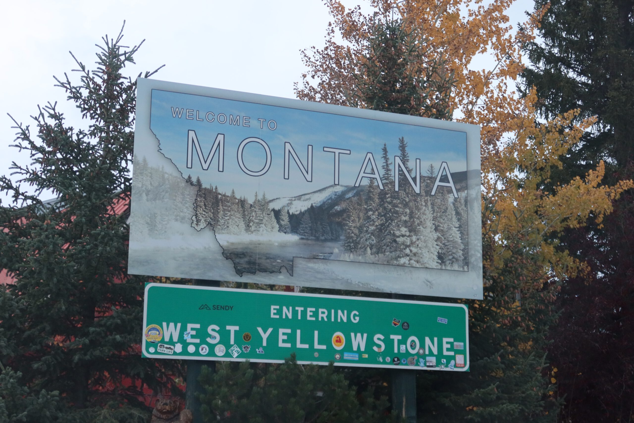 Welcome to Montana road sign at the West Yellowstone entrance, surrounded by pine trees and autumn foliage, marking the gateway to Yellowstone National Park.