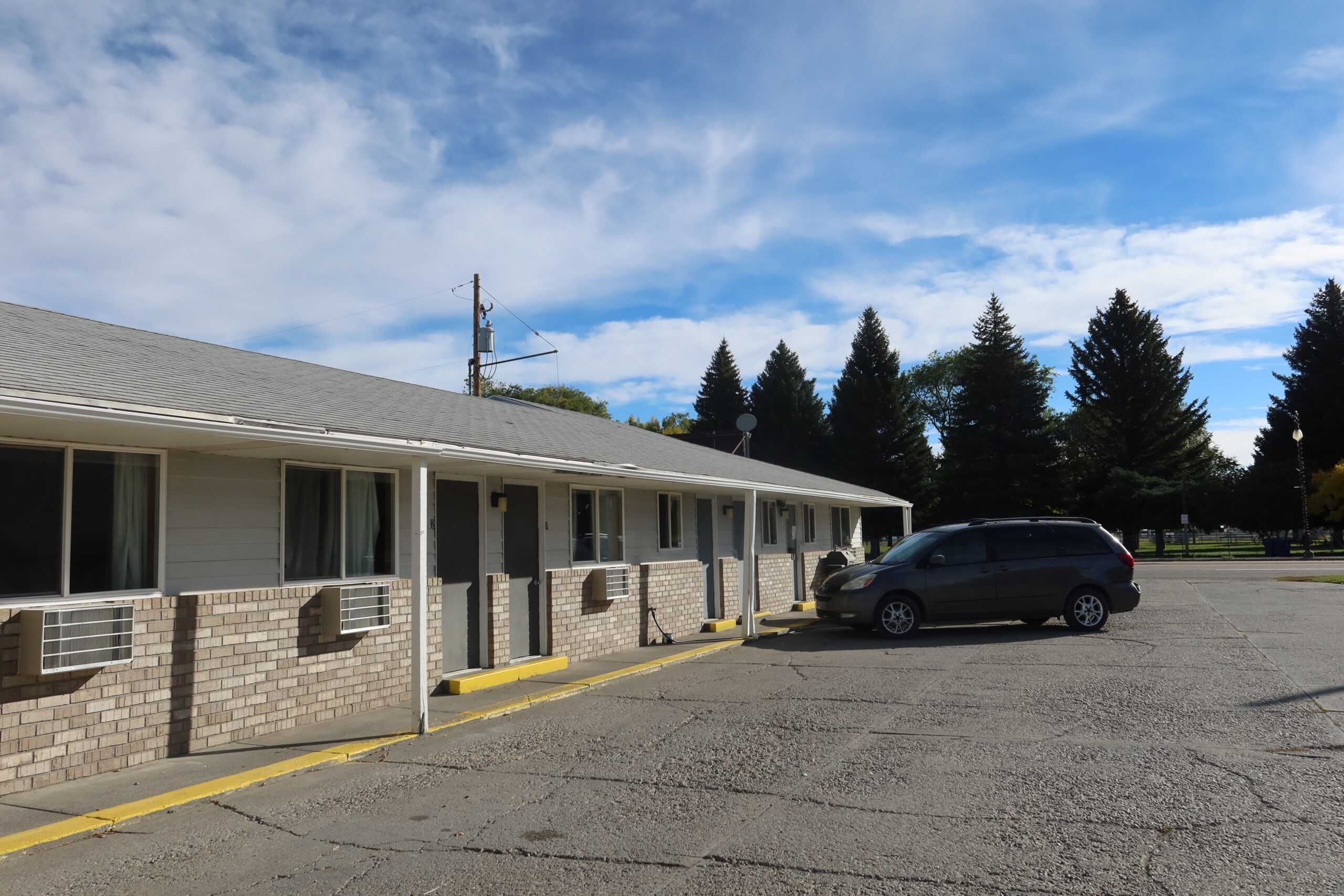 Exterior walkway of Arco Inn Motel in Arco, Idaho, showing a classic single story American roadside motel with room entrances, air conditioning units, on site parking, and wide open Idaho sky