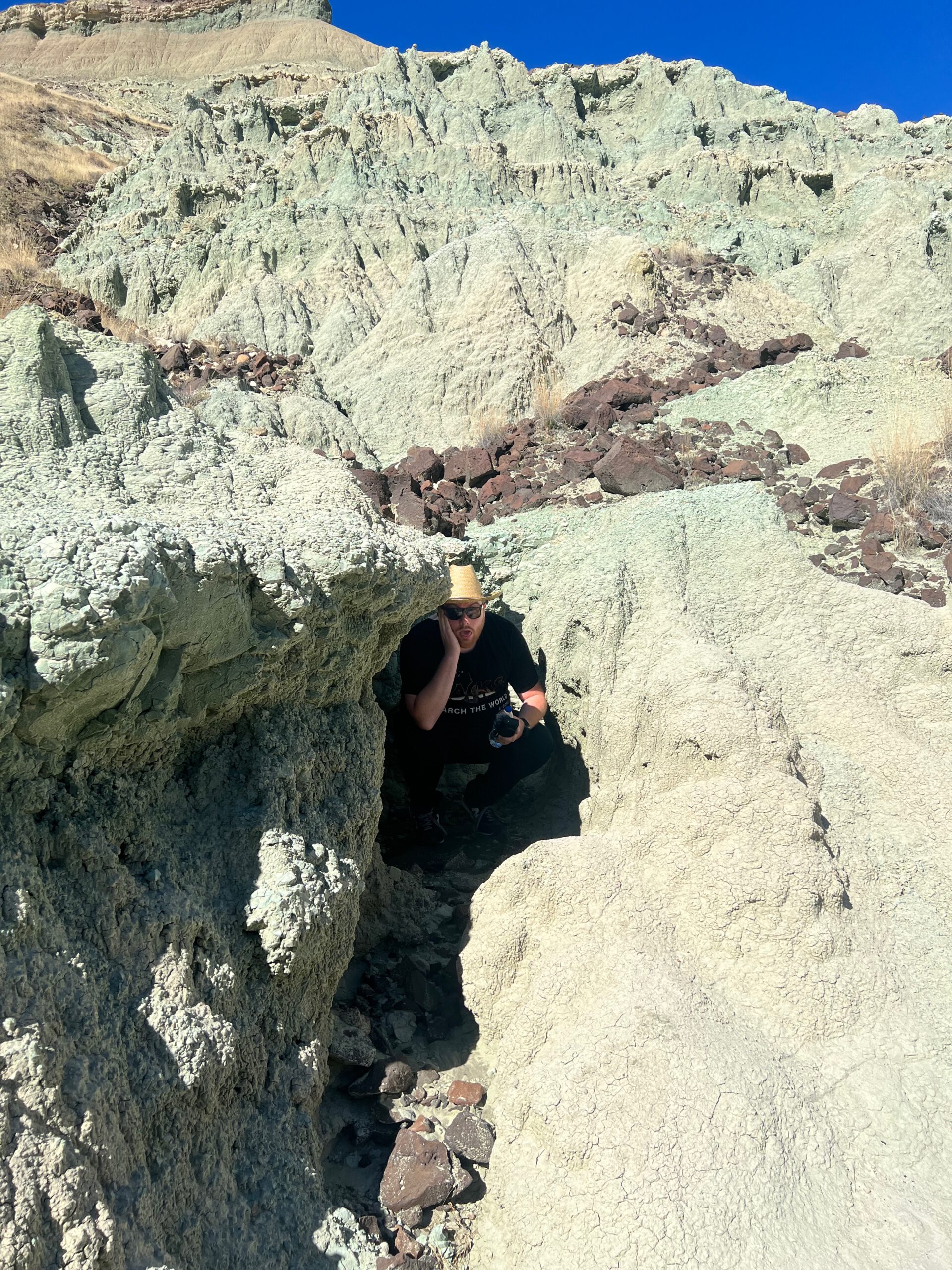 Timeuw, Timo van den Berg hiking a narrow trail at John Day Fossil Beds National Monument, standing between pale green clay formations and volcanic rock layers in Eastern Oregon