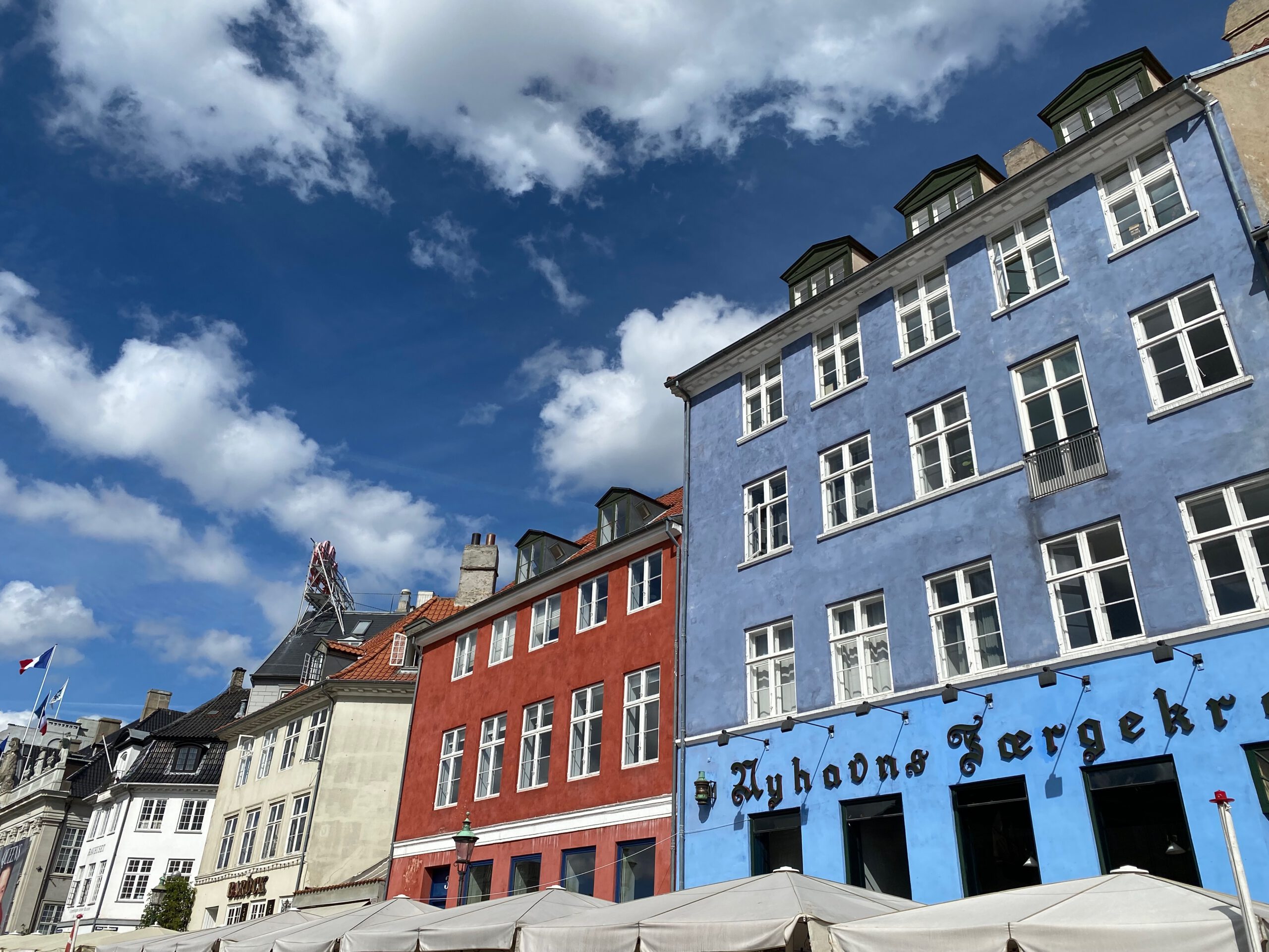 Nyhavn Copenhagen travel guide review, iconic colorful waterfront houses with café umbrellas and historic facades along Nyhavn harbor under a bright blue sky in Copenhagen.