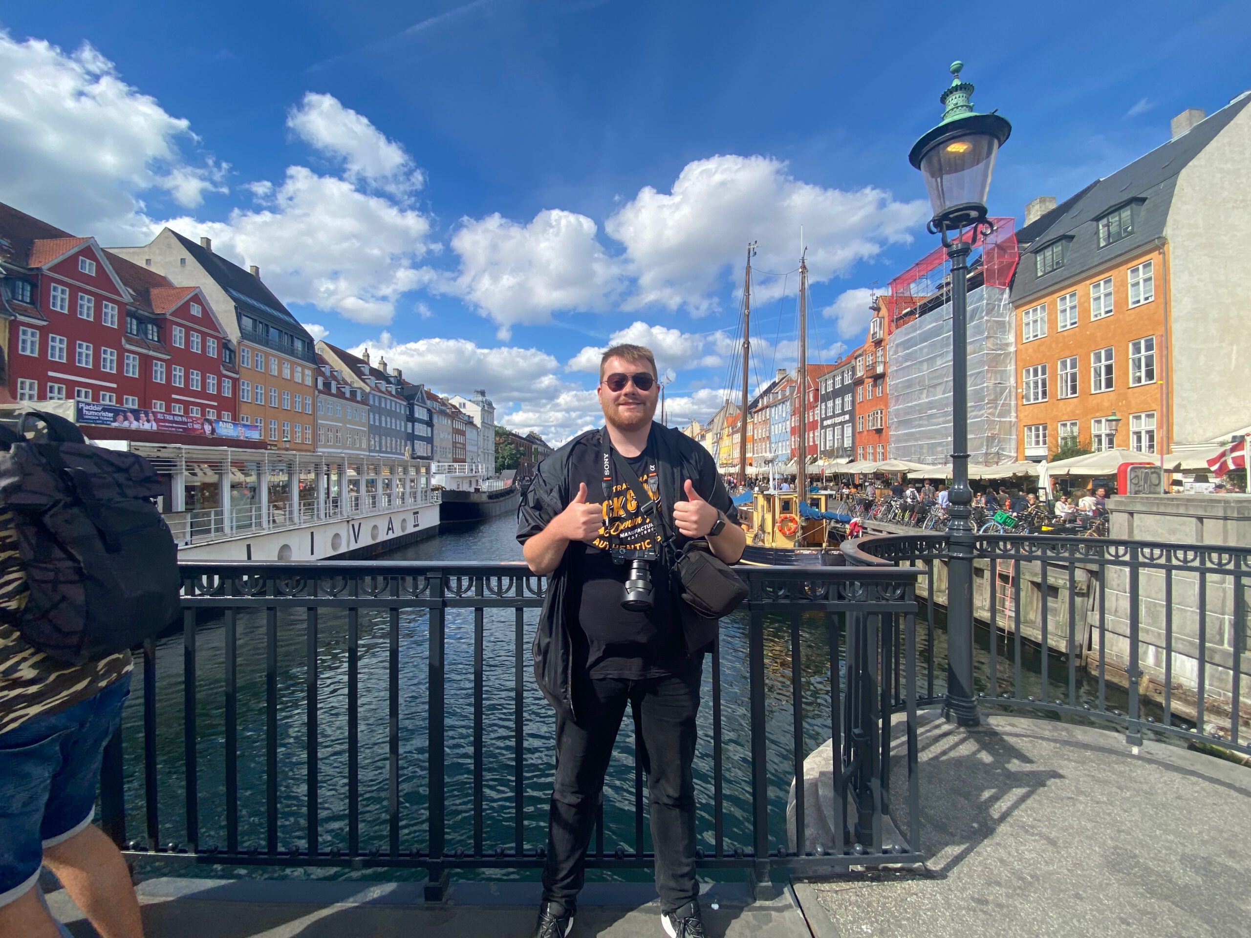 Nyhavn Copenhagen travel guide review, travel moment of Timo van den Berg at Nyhavn harbor featuring a visitor posing by the canal with classic wooden boats, vibrant houses, and blue skies in central Copenhagen.
